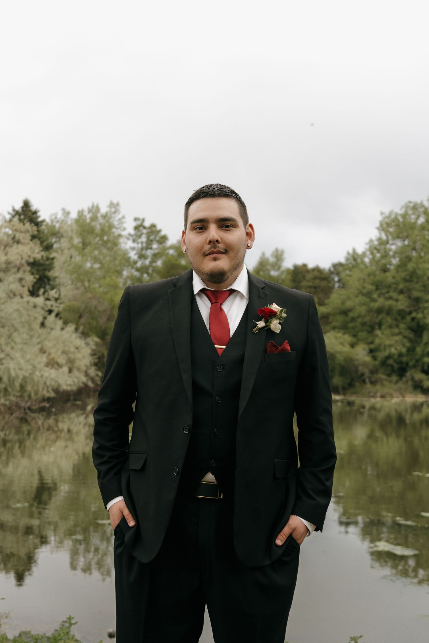 Groom in black three-piece suit with red tie and boutonniere standing by a pond with trees in background.