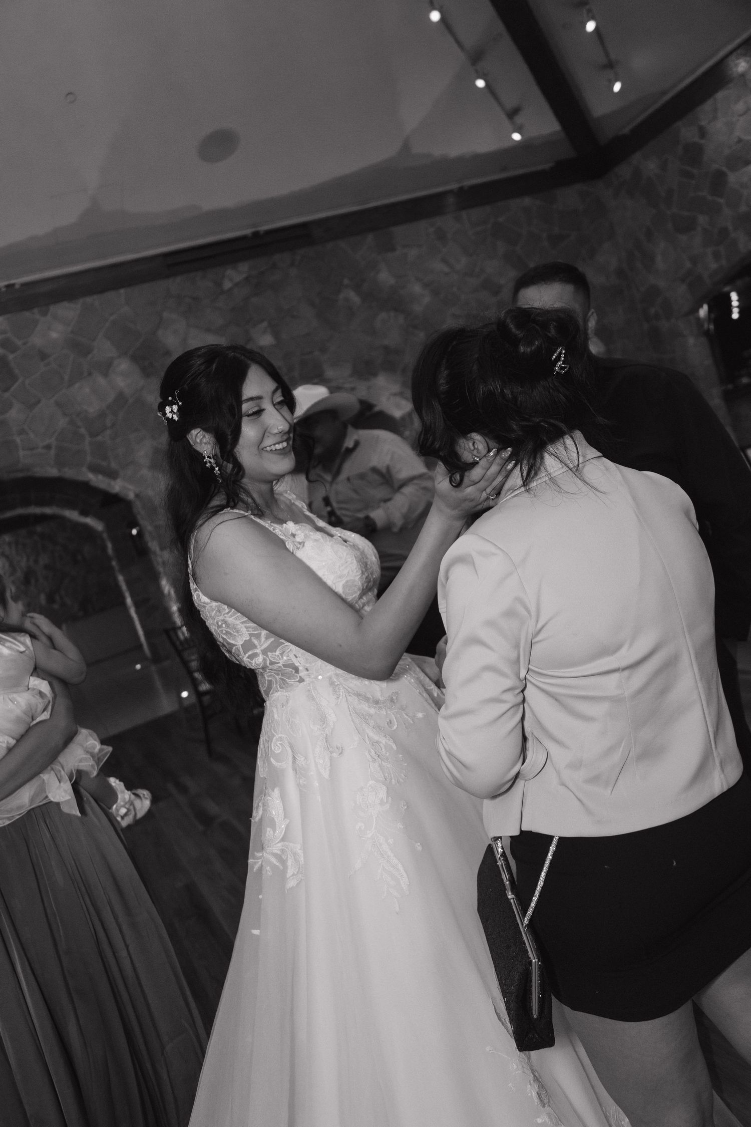 Black and white photo of smiling bride in lace gown dancing joyfully with guest at reception.
