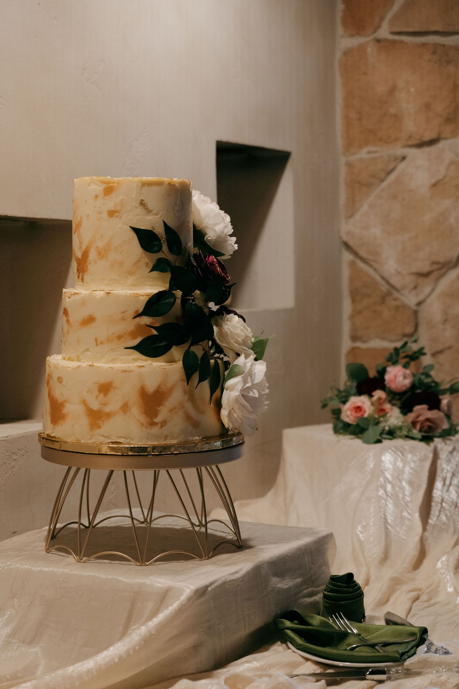 Elegant three-tier wedding cake with gold brush strokes and white floral arrangement on gold stand.