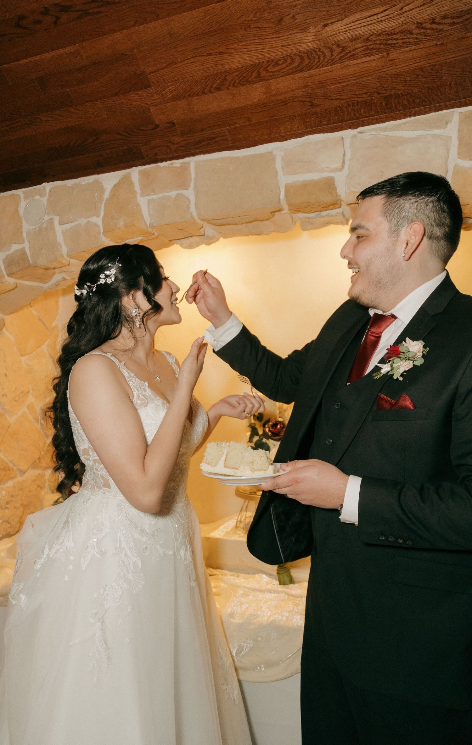 Bride and groom sharing wedding cake during reception near stone fireplace.