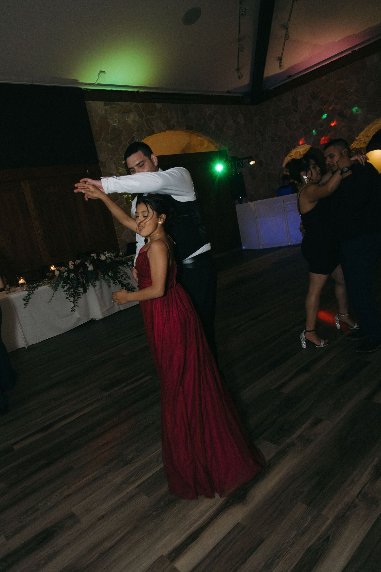 Bridesmaid in red gown being twirled by a groomsman on the colorfully lit wedding reception dance floor.