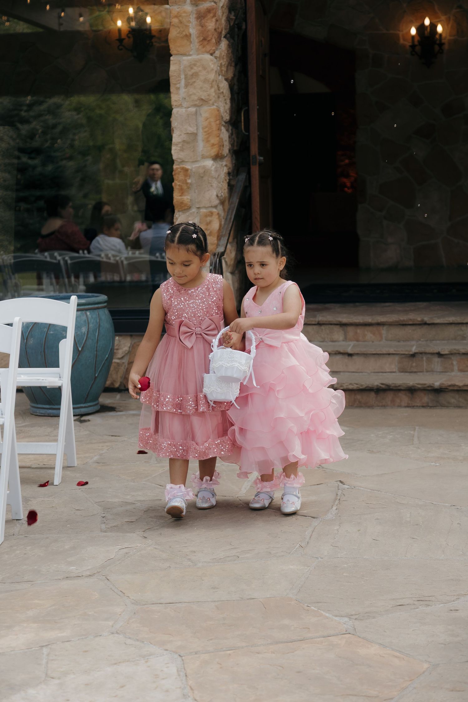 Two little flower girls in pink dresses walk down the aisle scattering rose petals at a wedding.