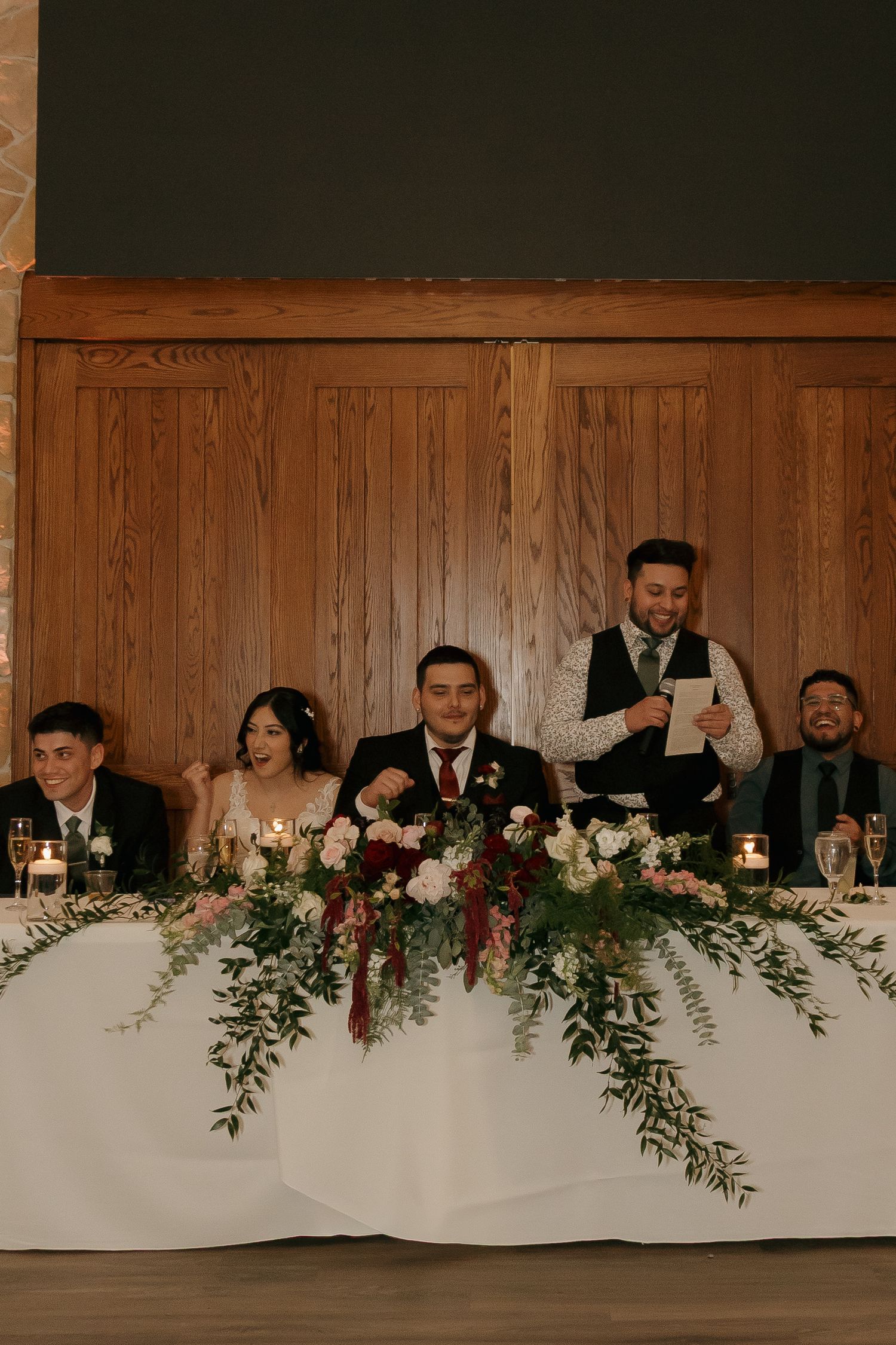 Wedding party seated at decorated head table with floral arrangements during reception speeches.