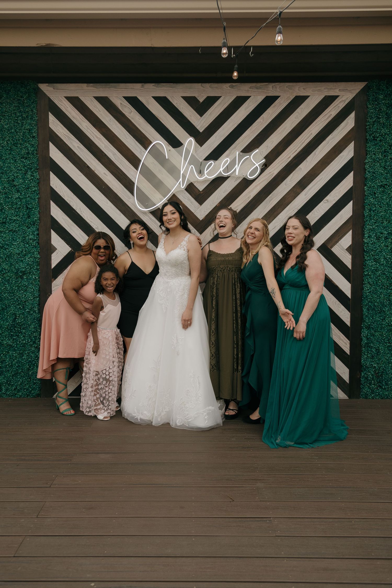 Bride in white gown poses with bridesmaids in green dresses before Cheers neon sign photo backdrop.