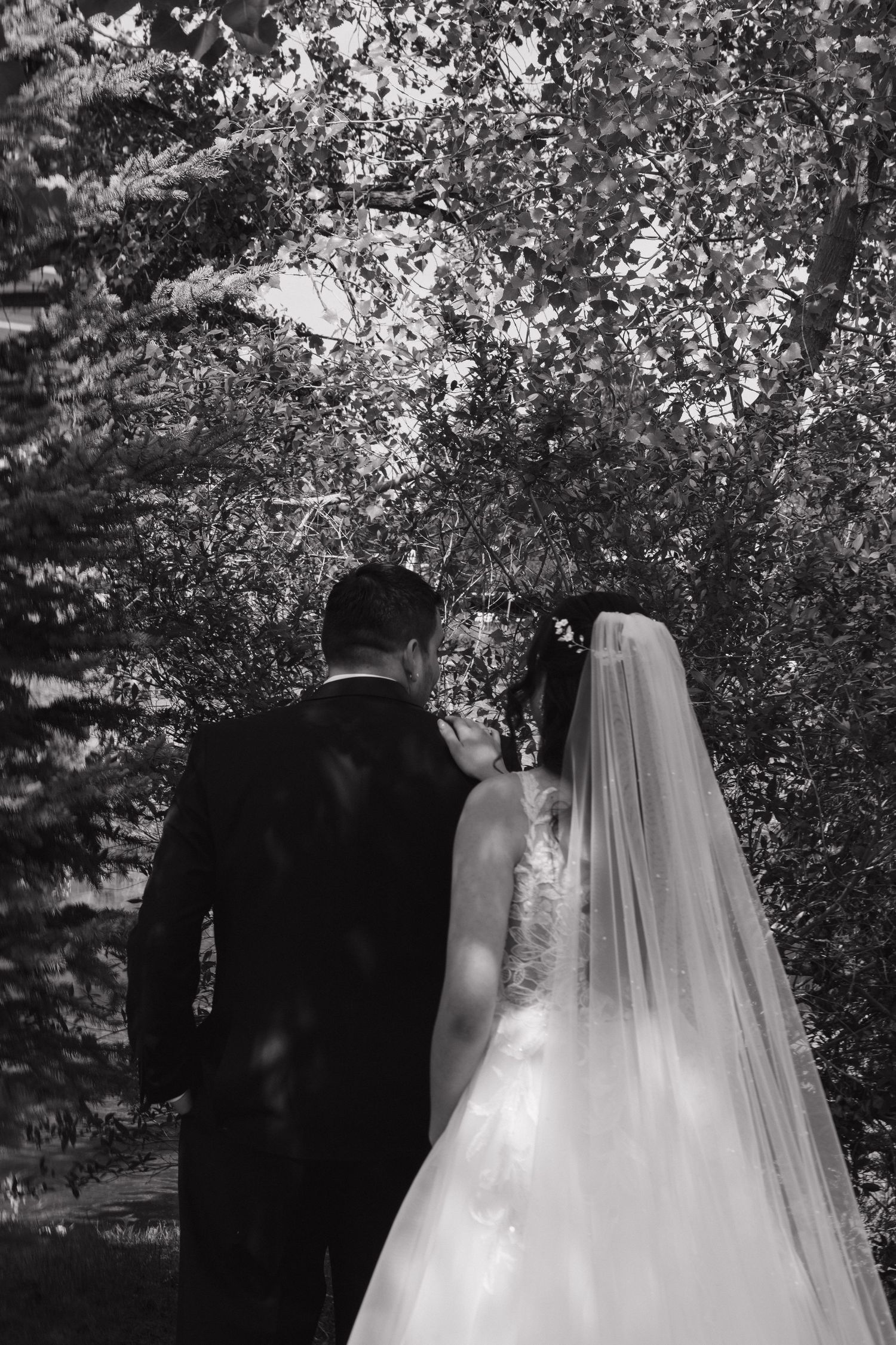 Black and white photo of bride and groom standing together beneath lush trees after outdoor wedding ceremony.