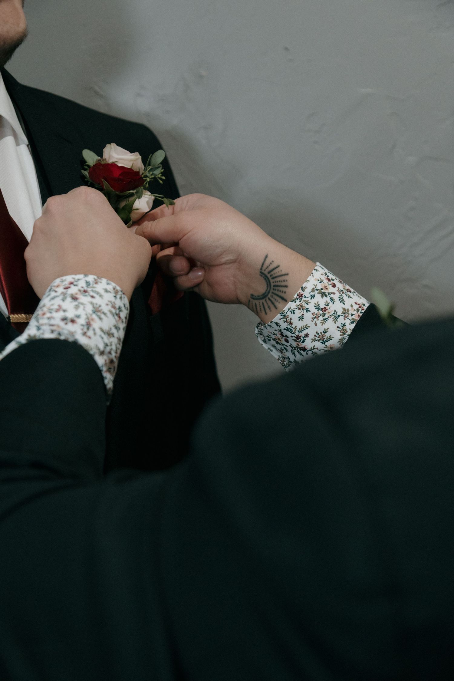 Tattooed hands pinning red and white boutonniere onto groom's dark suit lapel before wedding.