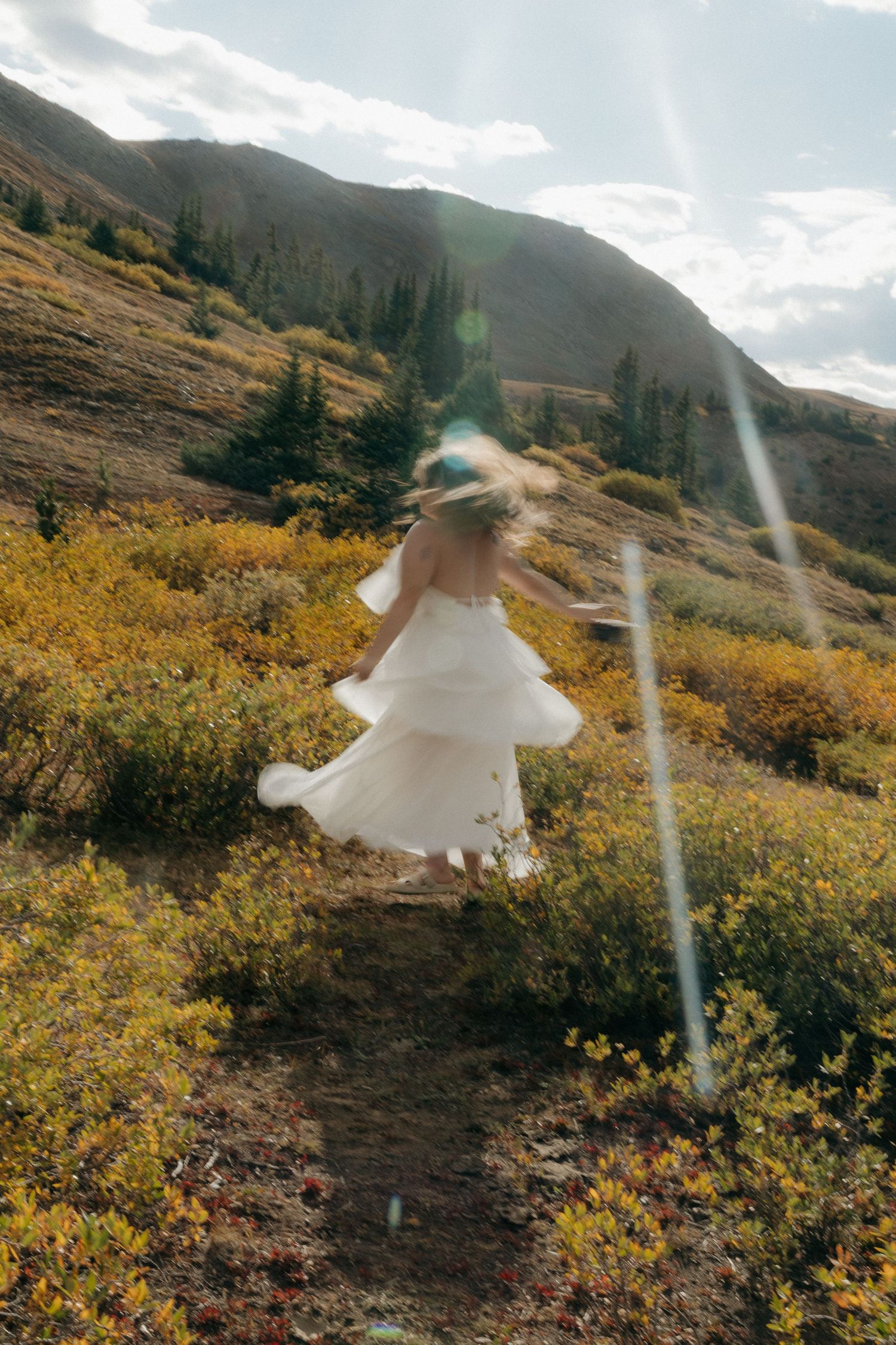 Bride in white dress running through golden autumn mountain meadow with lens flare and hills in background.