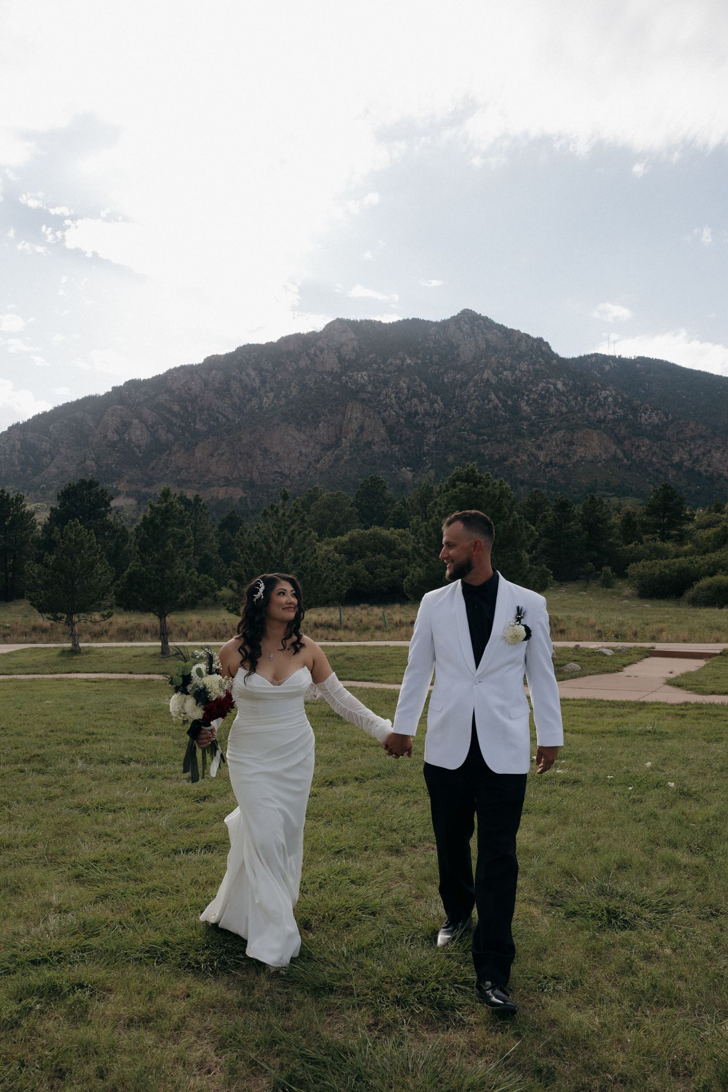 A bride and groom hold hands walking through a green meadow with a stunning rocky mountain backdrop on their wedding day.