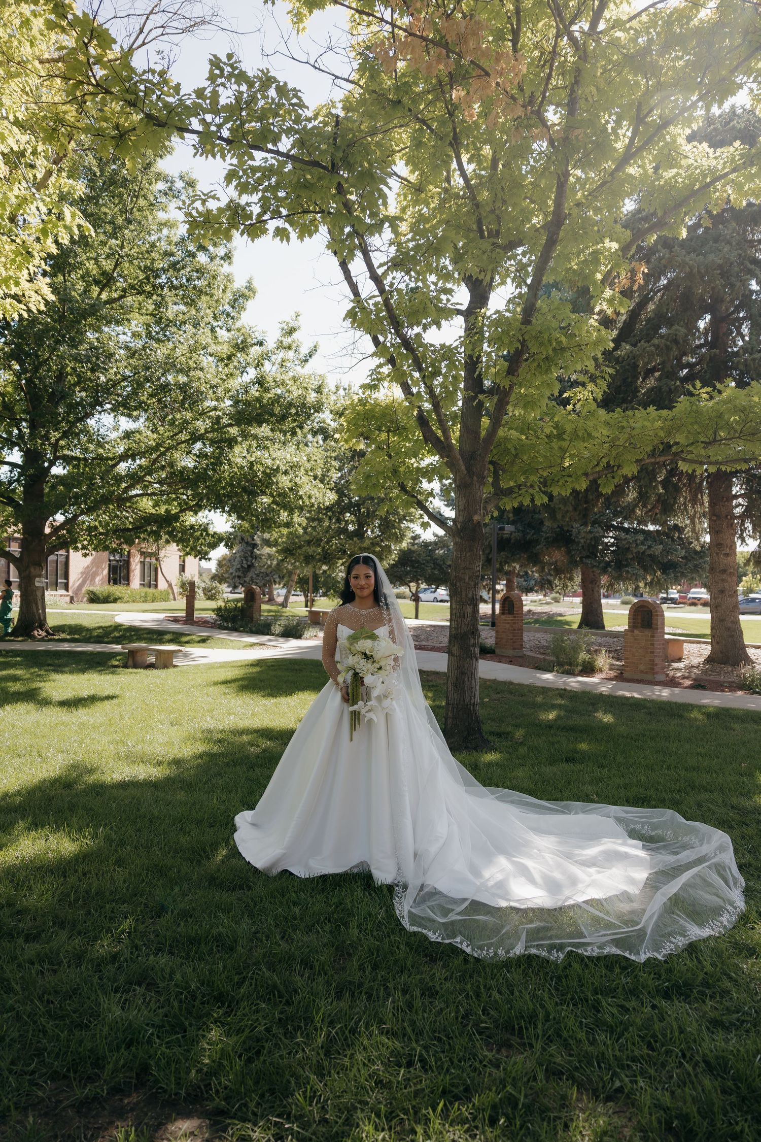 A bride in a flowing white gown with a long veil holds a bouquet while standing in a lush green park.