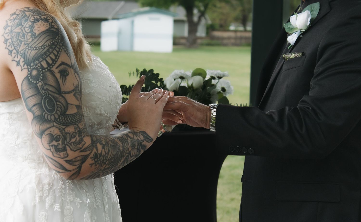 Two people in formal wedding attire stand close together during an outdoor ceremony.