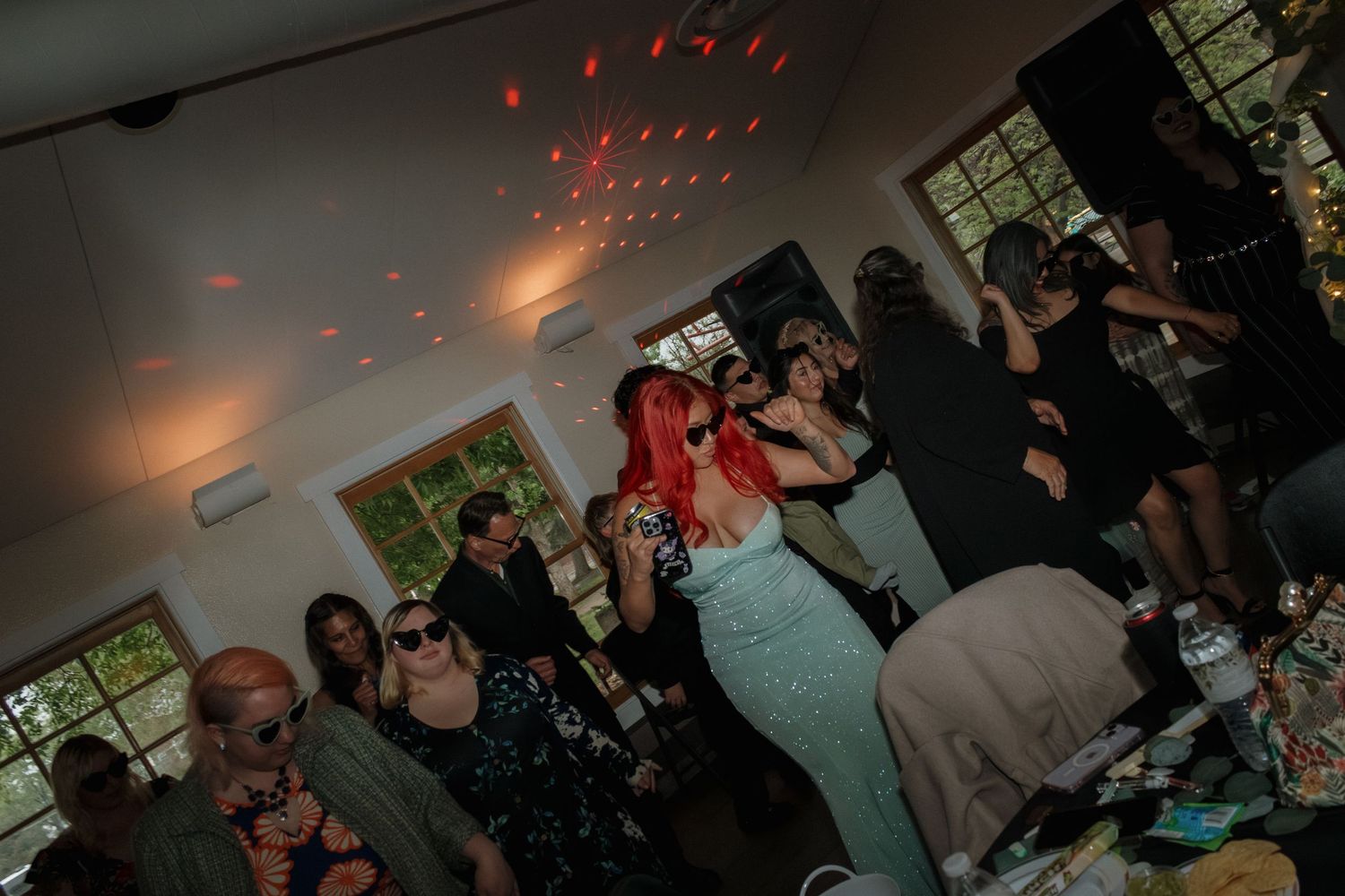 Party guests dancing under red disco lights in a dimly lit room with windows.