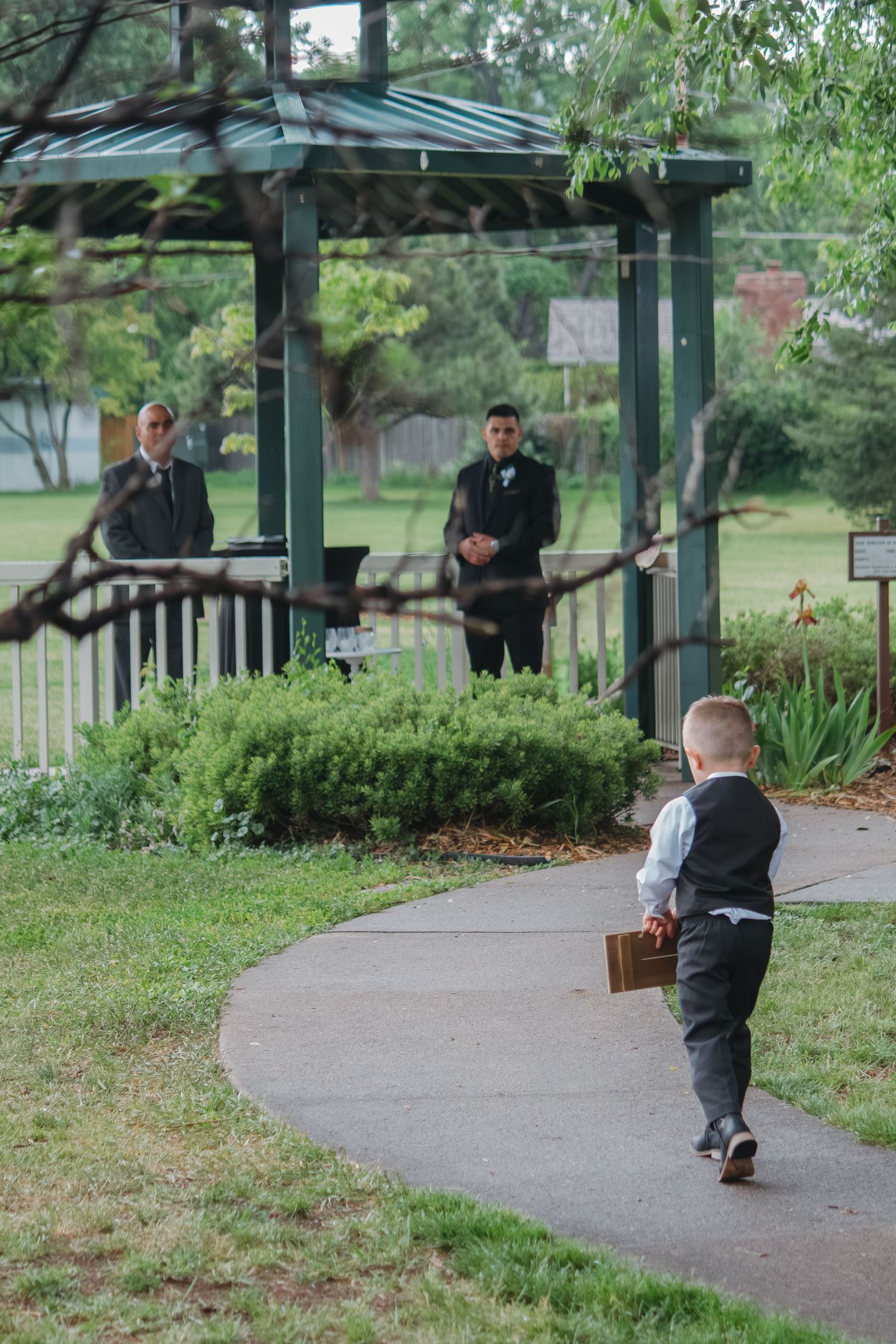 A small child in formal wear walks along a path toward a covered gazebo with greenery.