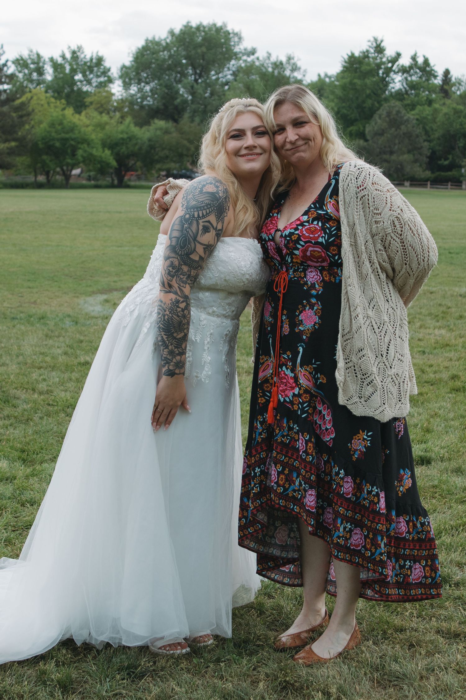 Outdoor wedding photo showing bride in white dress with tattoos alongside guest in floral dress.