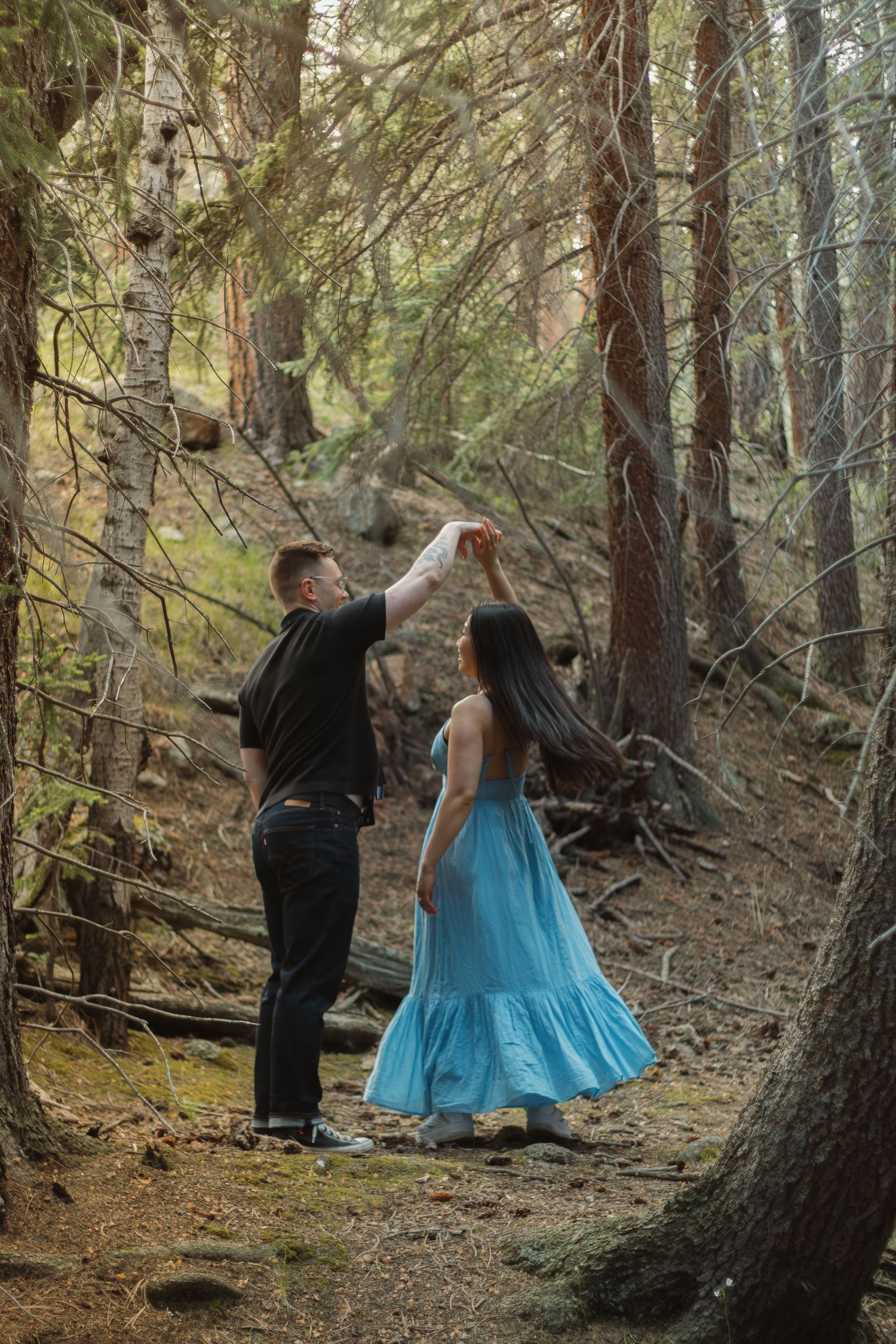 Two people dressed in black and blue stand together amid tall trees in a forest setting.