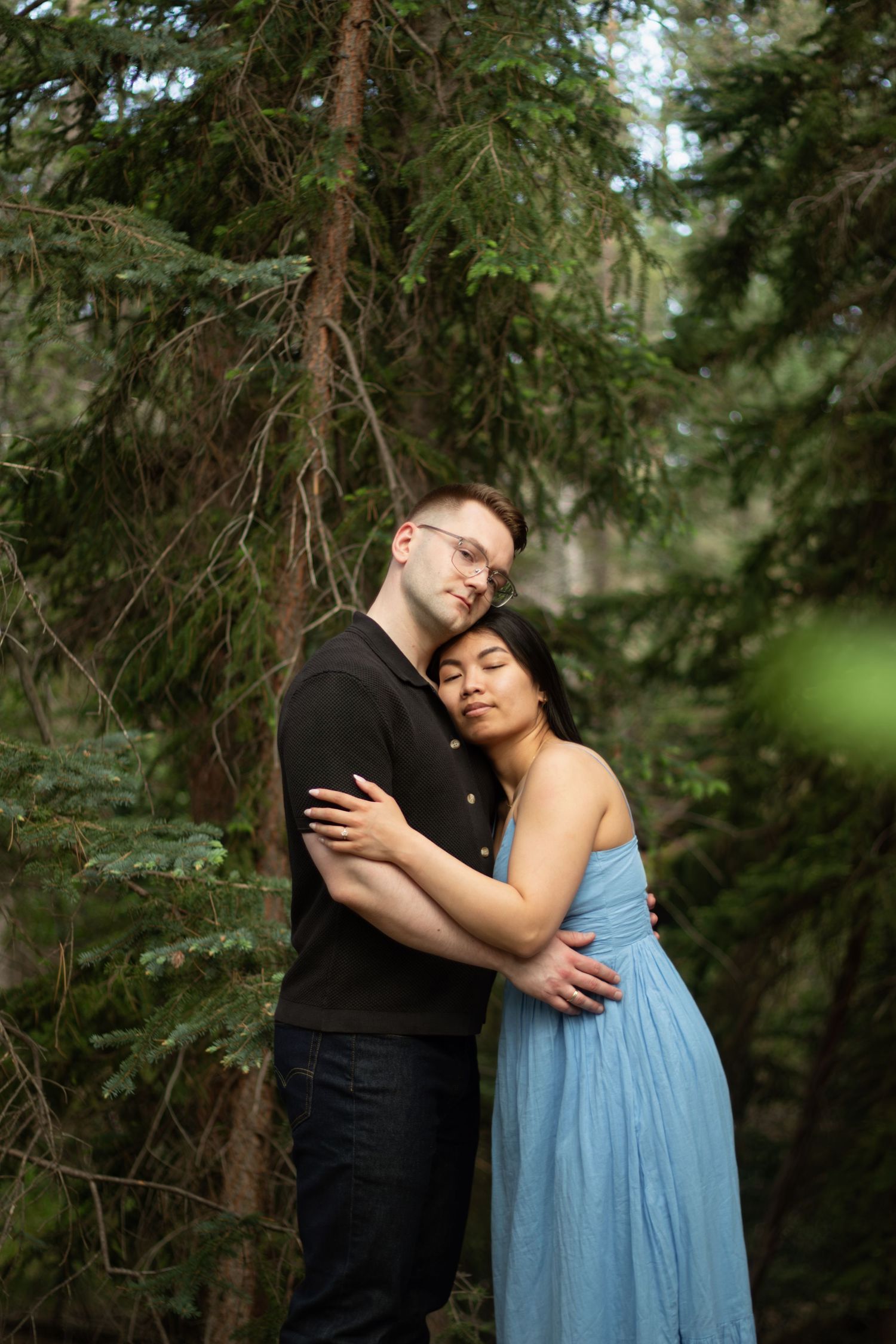A couple shares an intimate moment in a forest, with her in a flowing blue dress and him in black.