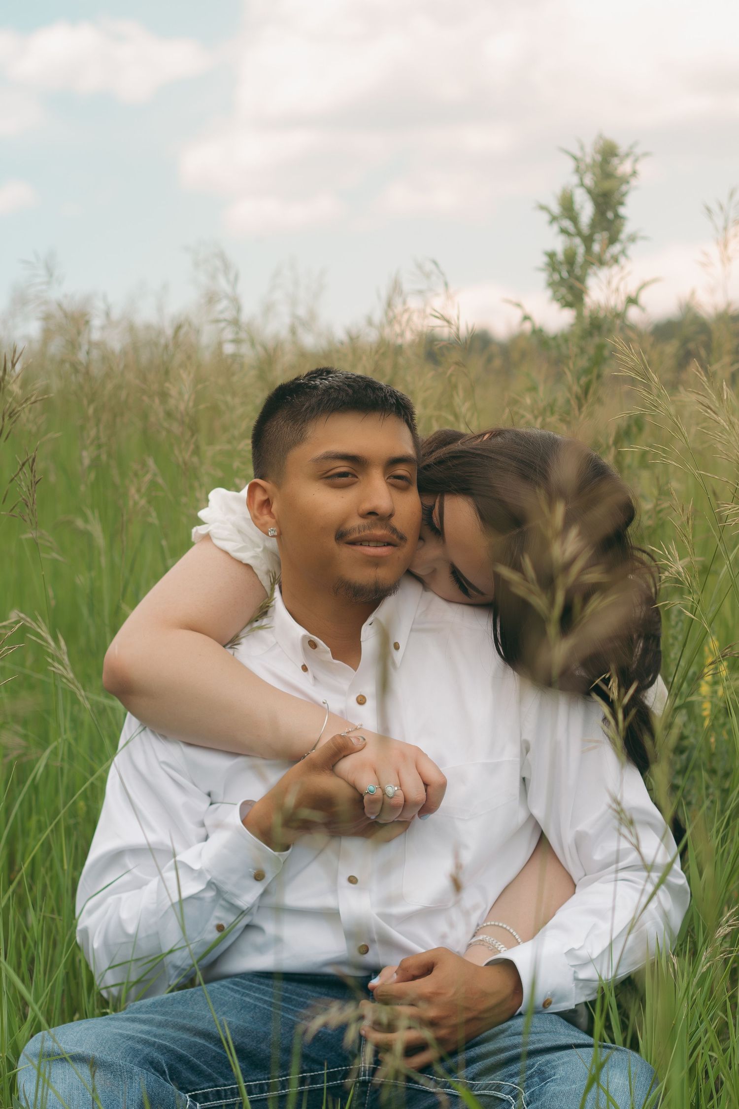 Couple in white shirts sharing tender moment while sitting in tall summer grass.