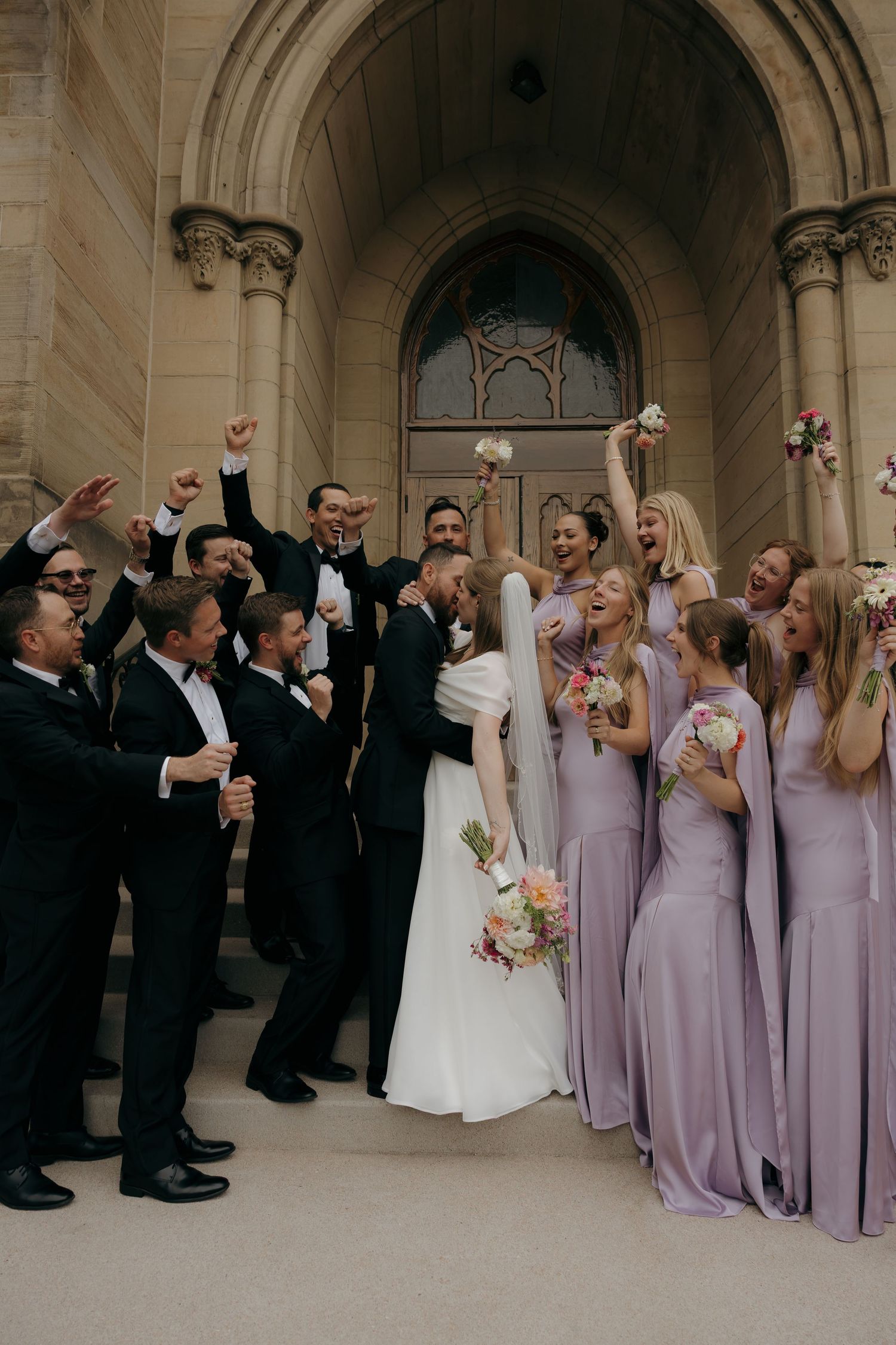 Joyful wedding party celebrates on church steps, bridesmaids in lavender gowns and groomsmen in black tuxedos cheering newlyweds.