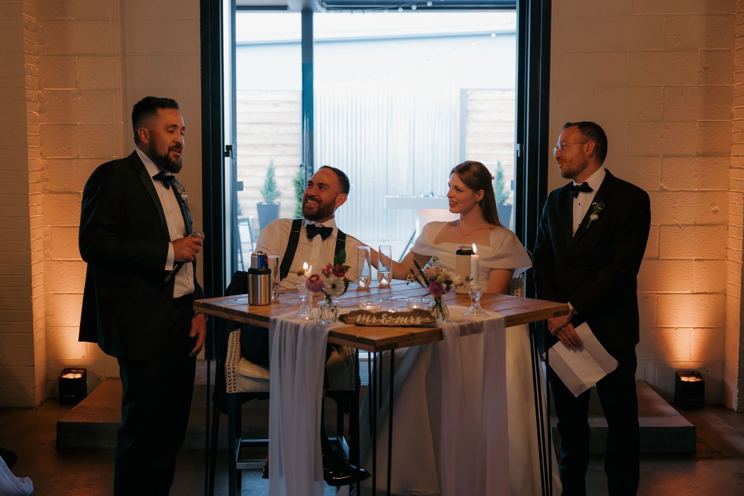 Wedding couple seated at sweetheart table while groomsmen give speeches in elegant venue with warm lighting.