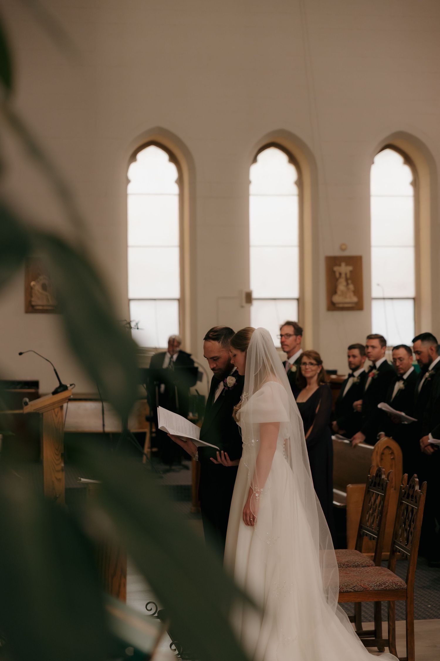 Bride in white veil and groom reading vows at church altar with arched windows and wedding party behind them.