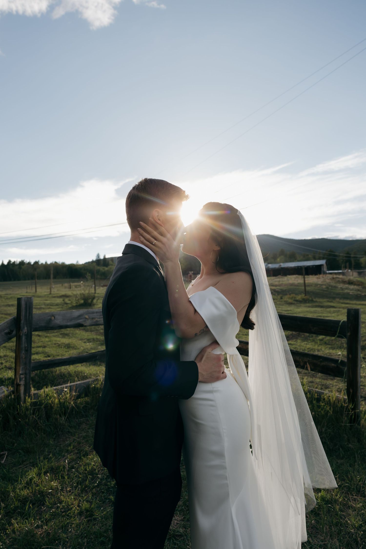 Bride and groom share a romantic kiss backlit by golden sun on a rustic farm, bride wearing white gown and veil.