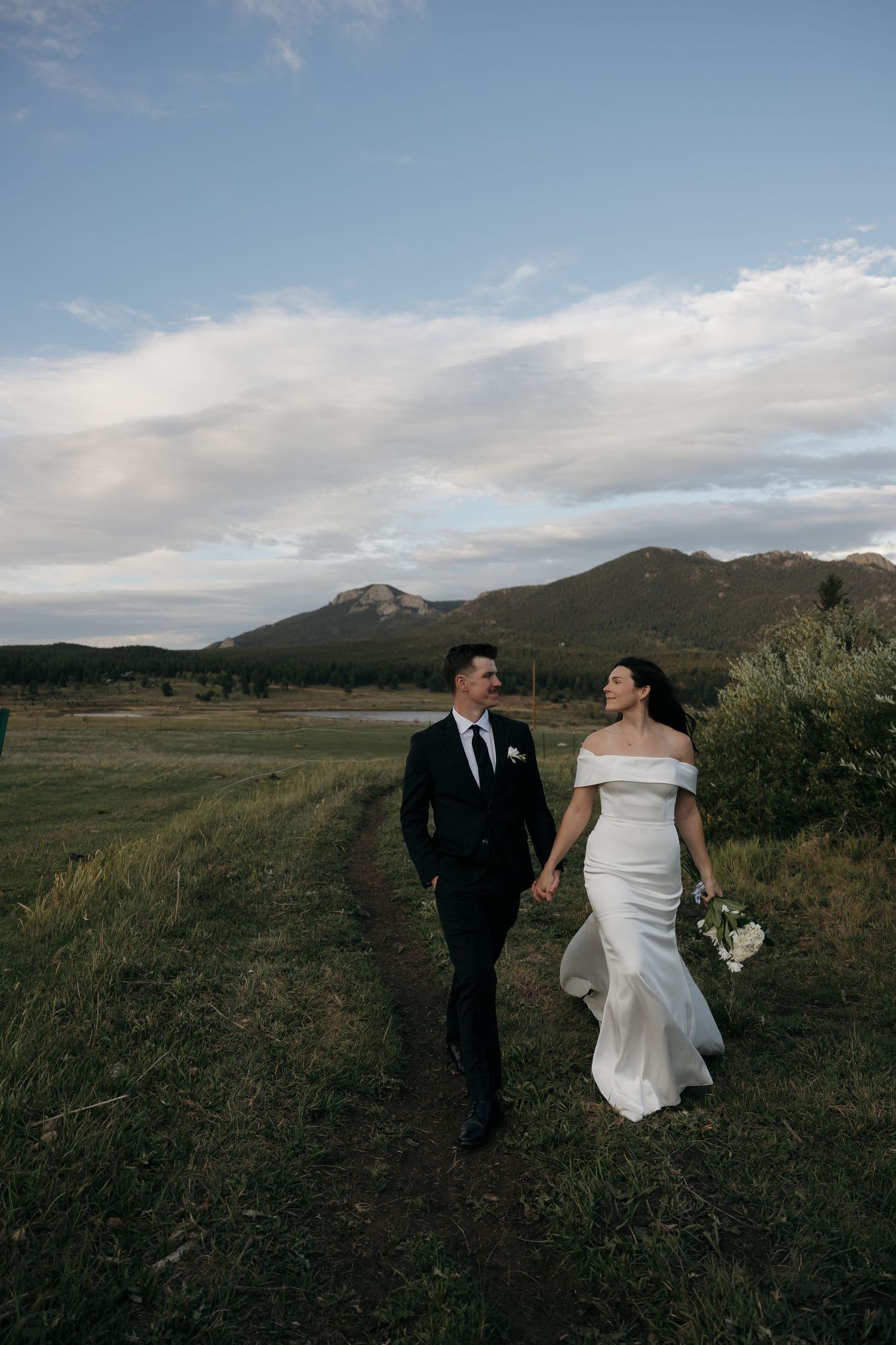 Bride and groom walking hand in hand along mountain meadow path at golden hour wedding.
