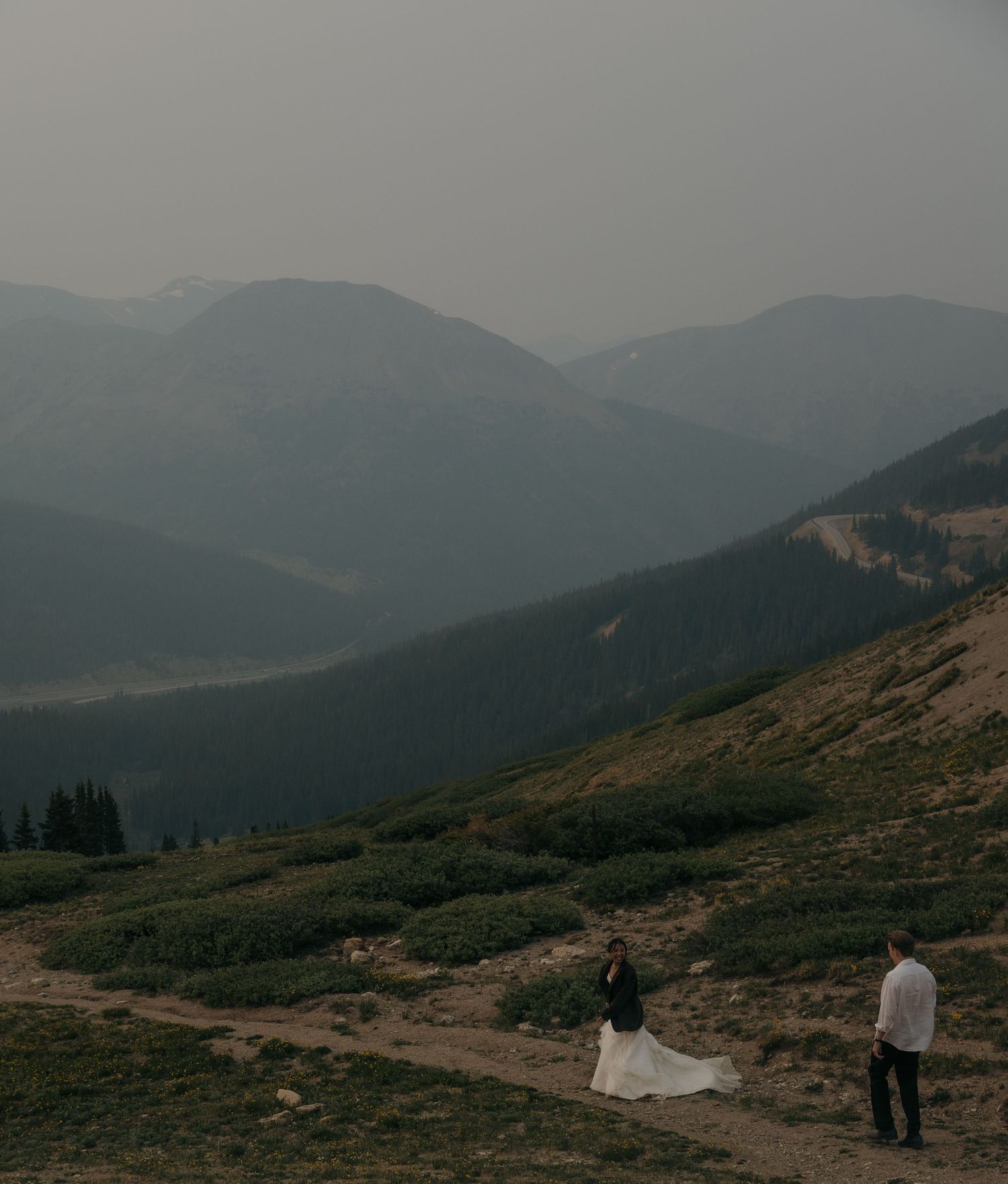 Distant view of bride and groom on a dramatic misty mountain trail with sweeping valley views.