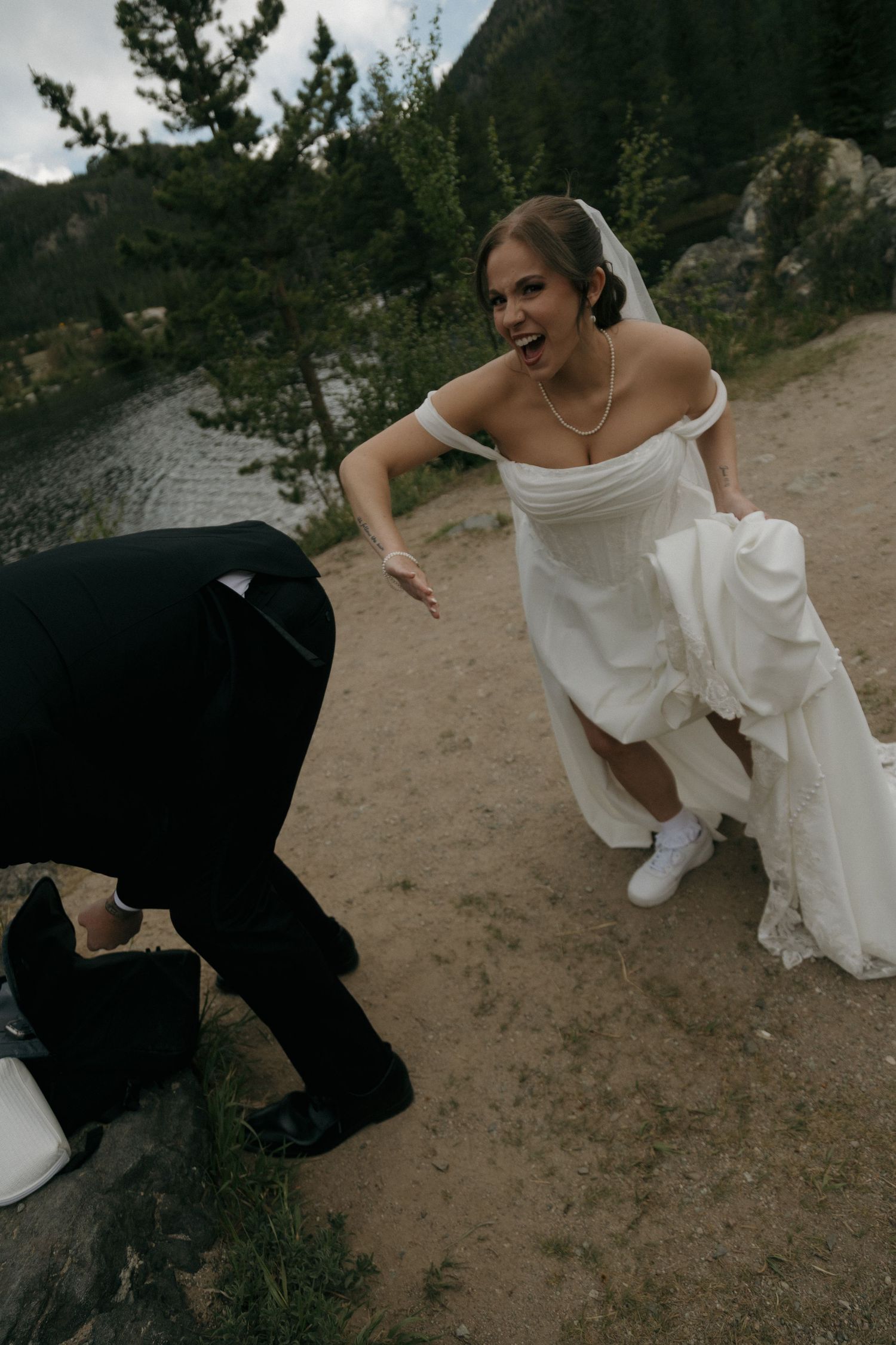 Bride in white gown laughing as groom kneels on rocky mountain path near a lake.