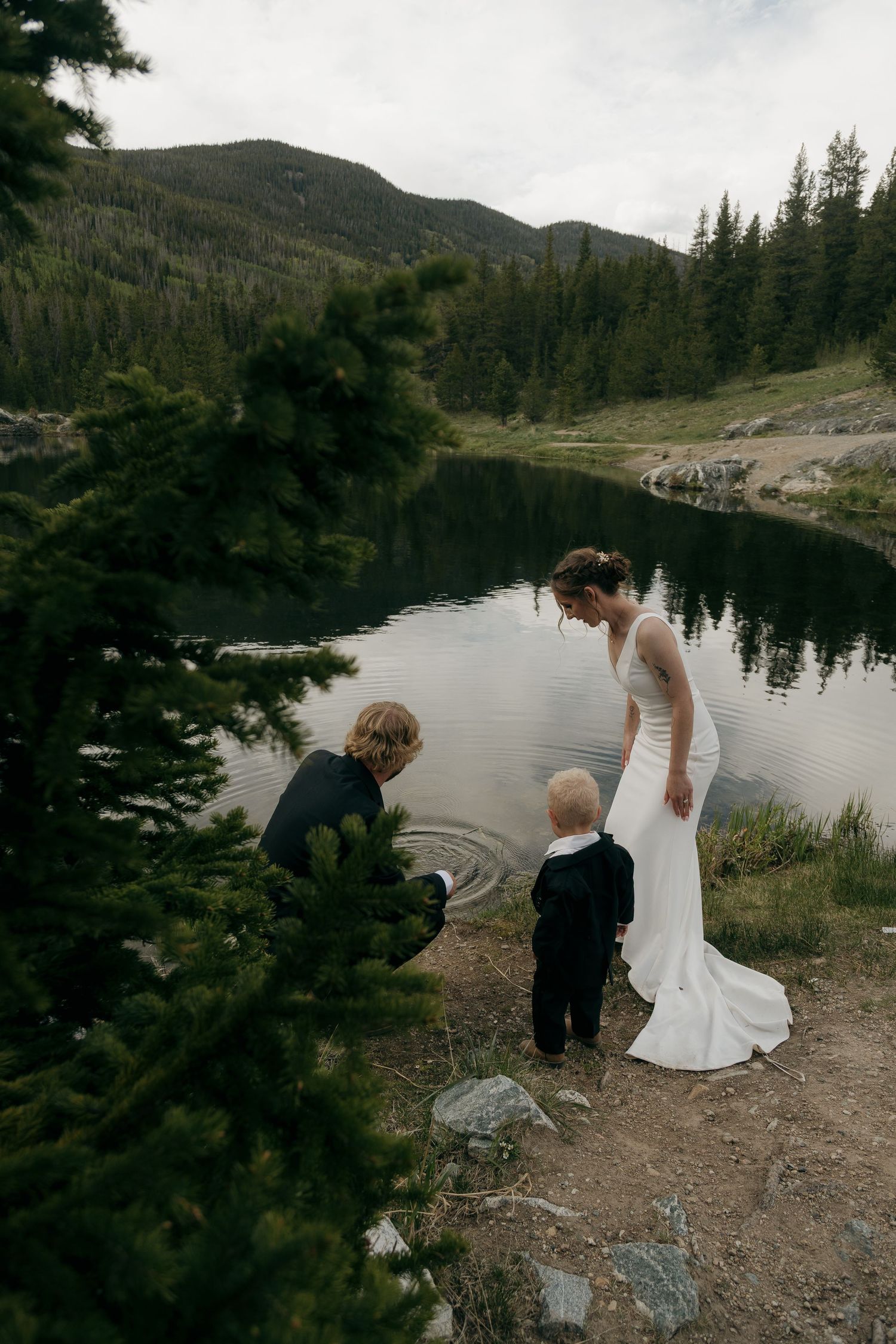 Bride in white gown watches two children play by a serene mountain lake.