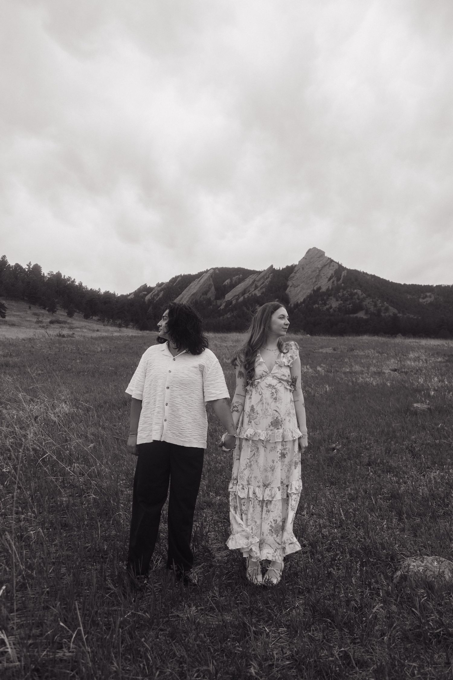 Black and white photo of two women holding hands walking through a field with mountain flatirons backdrop.