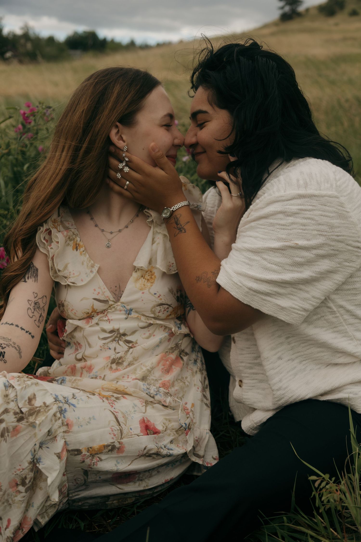 Two women share a romantic moment in a wildflower field, one cradling the other's face as they lean in to kiss.