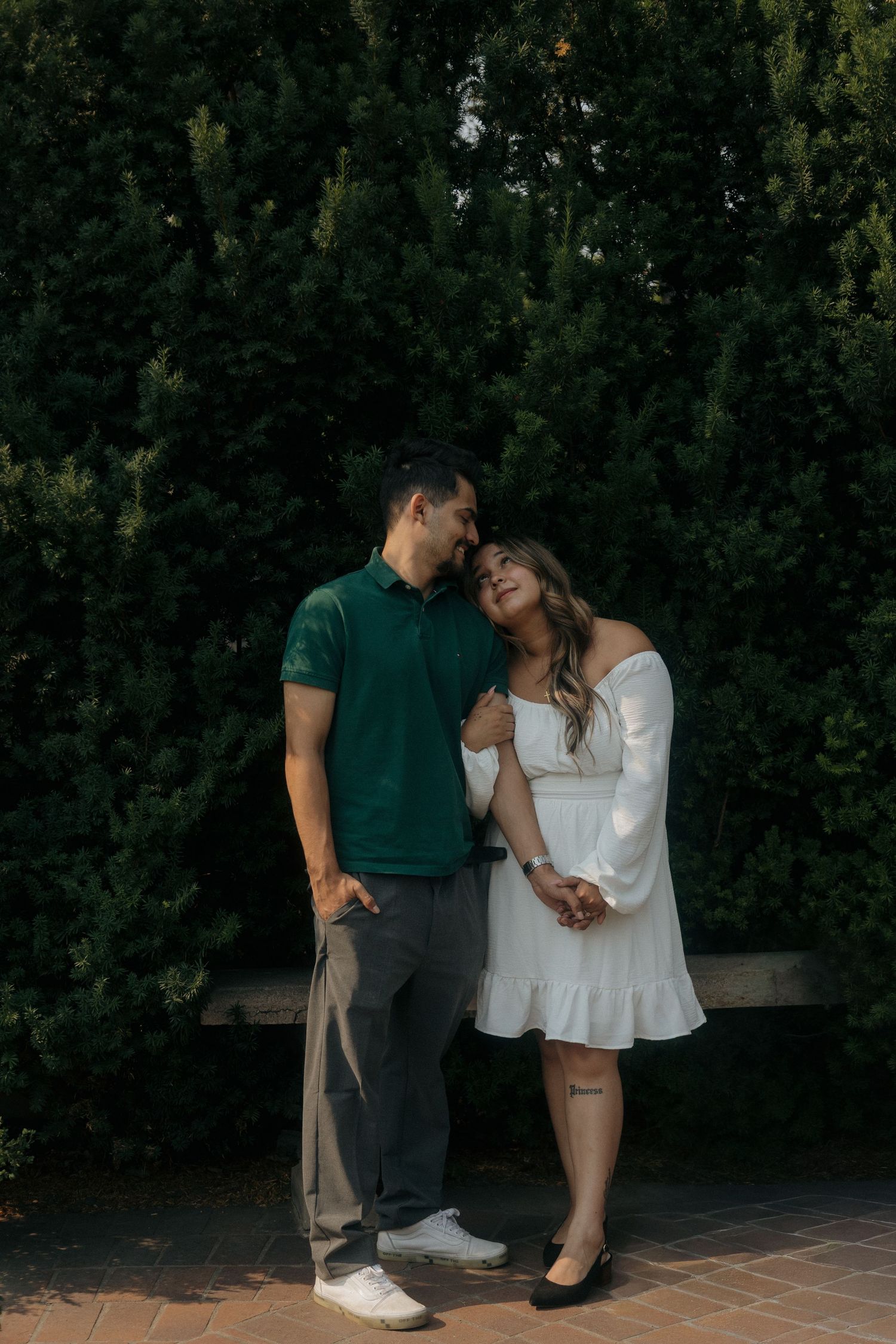 Couple embracing outdoors before tall evergreen hedges, man in green polo and woman in white dress.