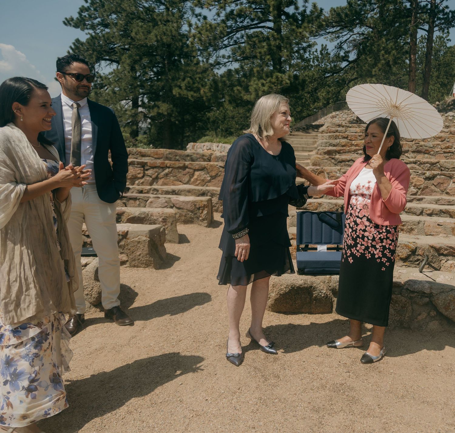 Wedding guests gather outdoors at a rocky mountain venue, bride holds white parasol umbrella.