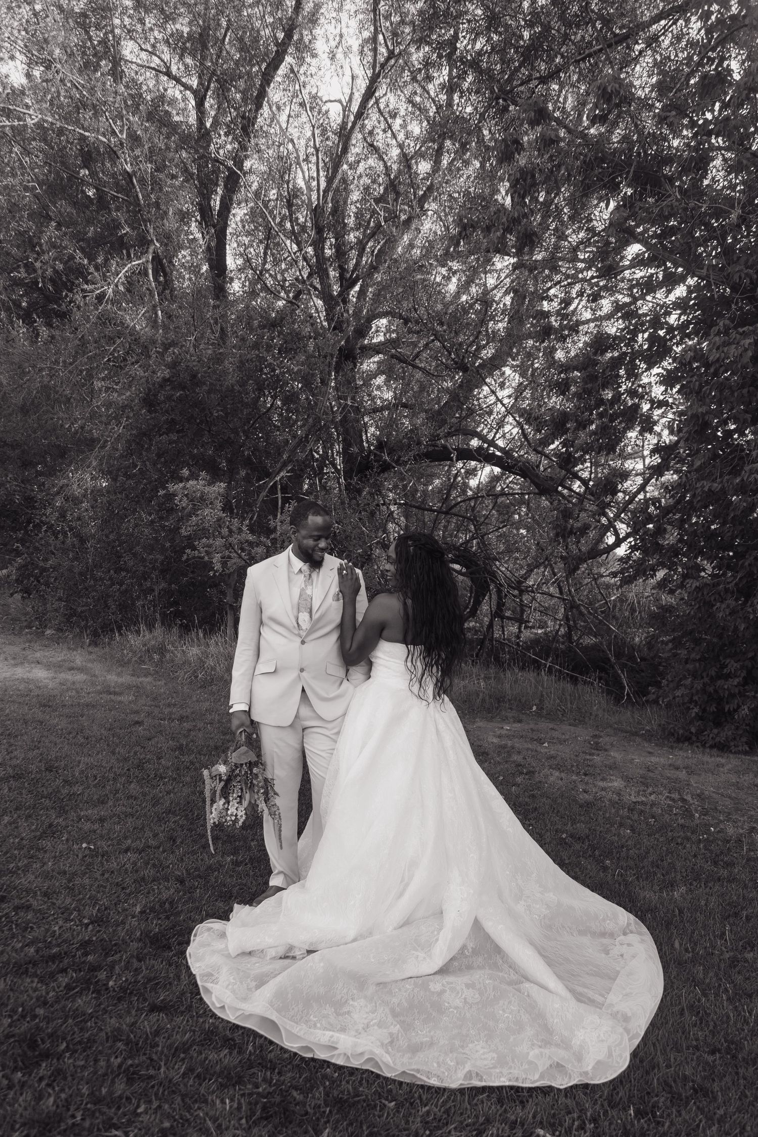 Black and white photo of bride and groom embracing near trees, showcasing the bride's elegant long train.