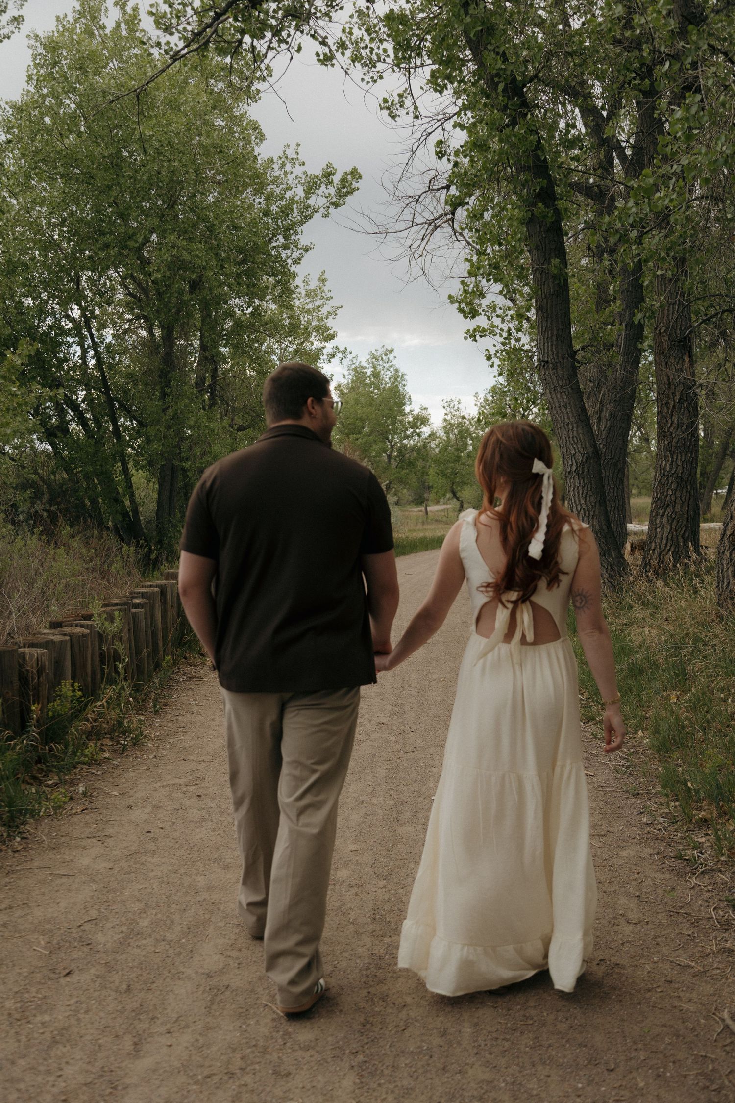 Couple holding hands walking down a tree-lined dirt path, woman in white dress and man in black shirt.