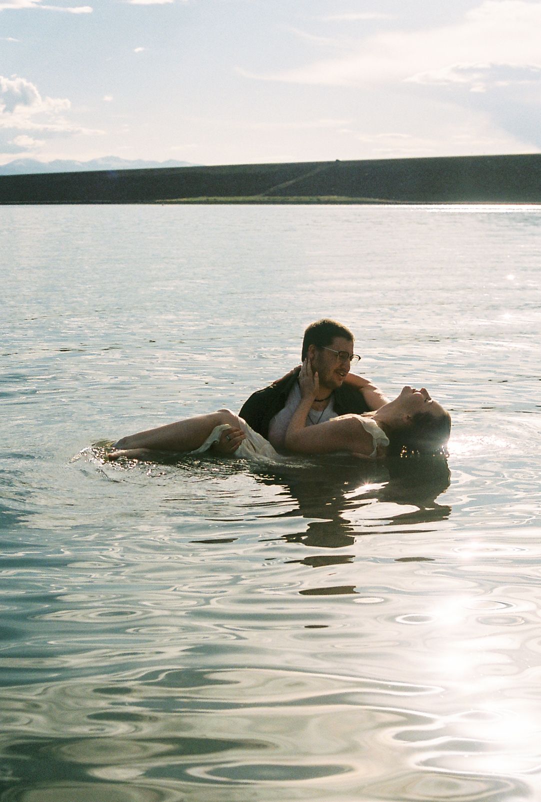 Couple floating together in calm lake water with scenic hills in the background.