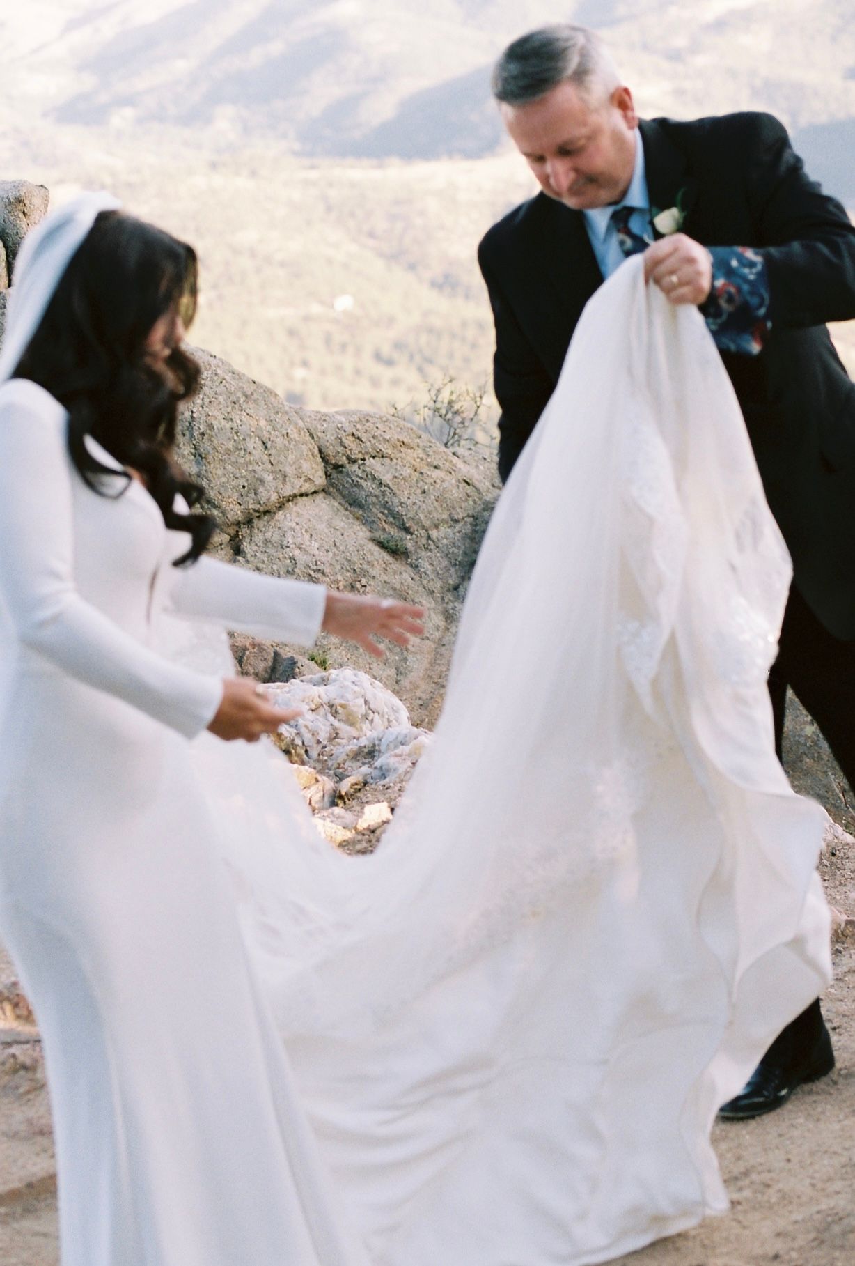 Father helps arrange bride's flowing white cathedral veil on a rocky mountain overlook before ceremony.