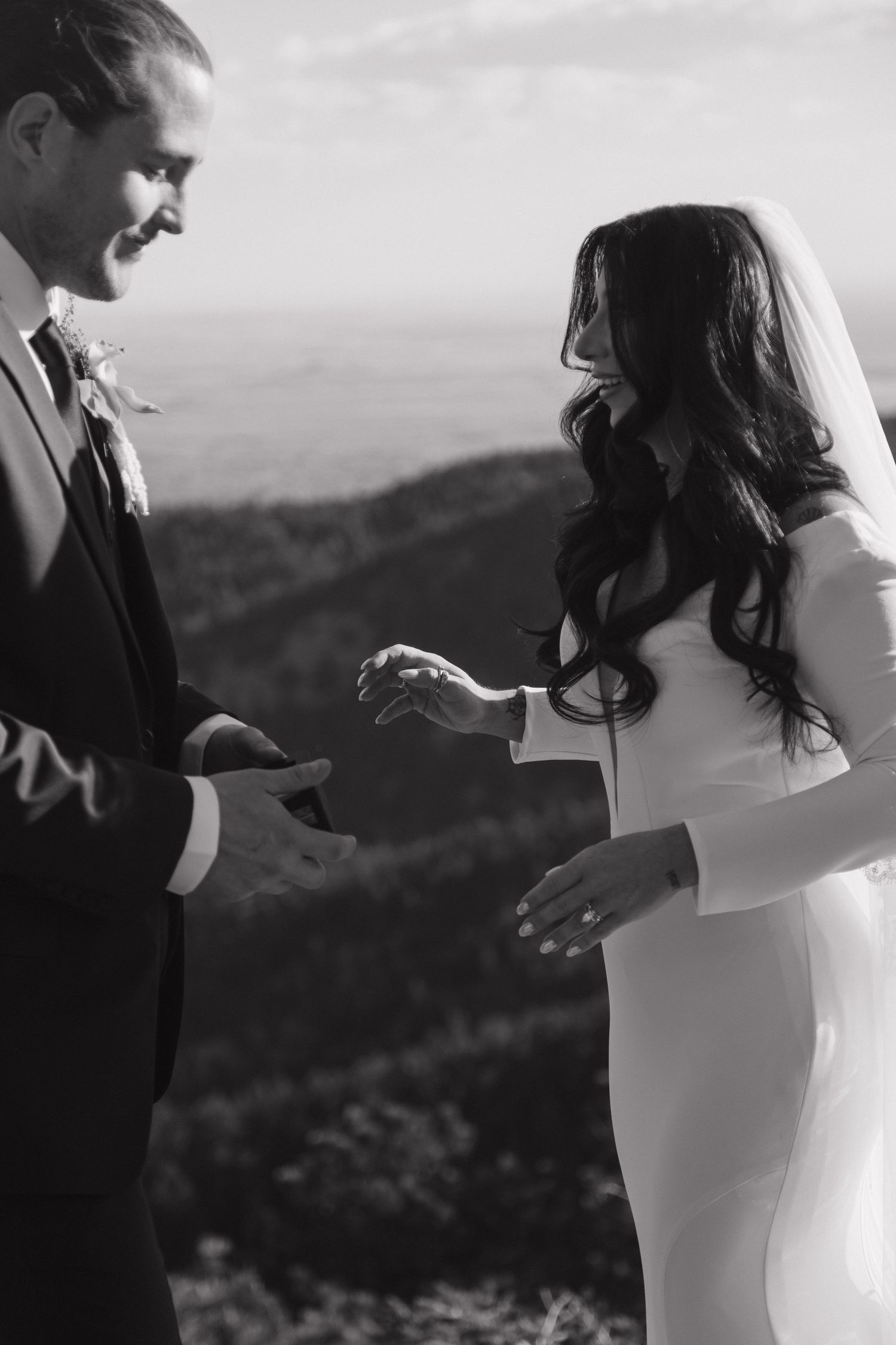 Black and white photo of smiling bride and groom exchanging rings on a mountain top during their elopement.