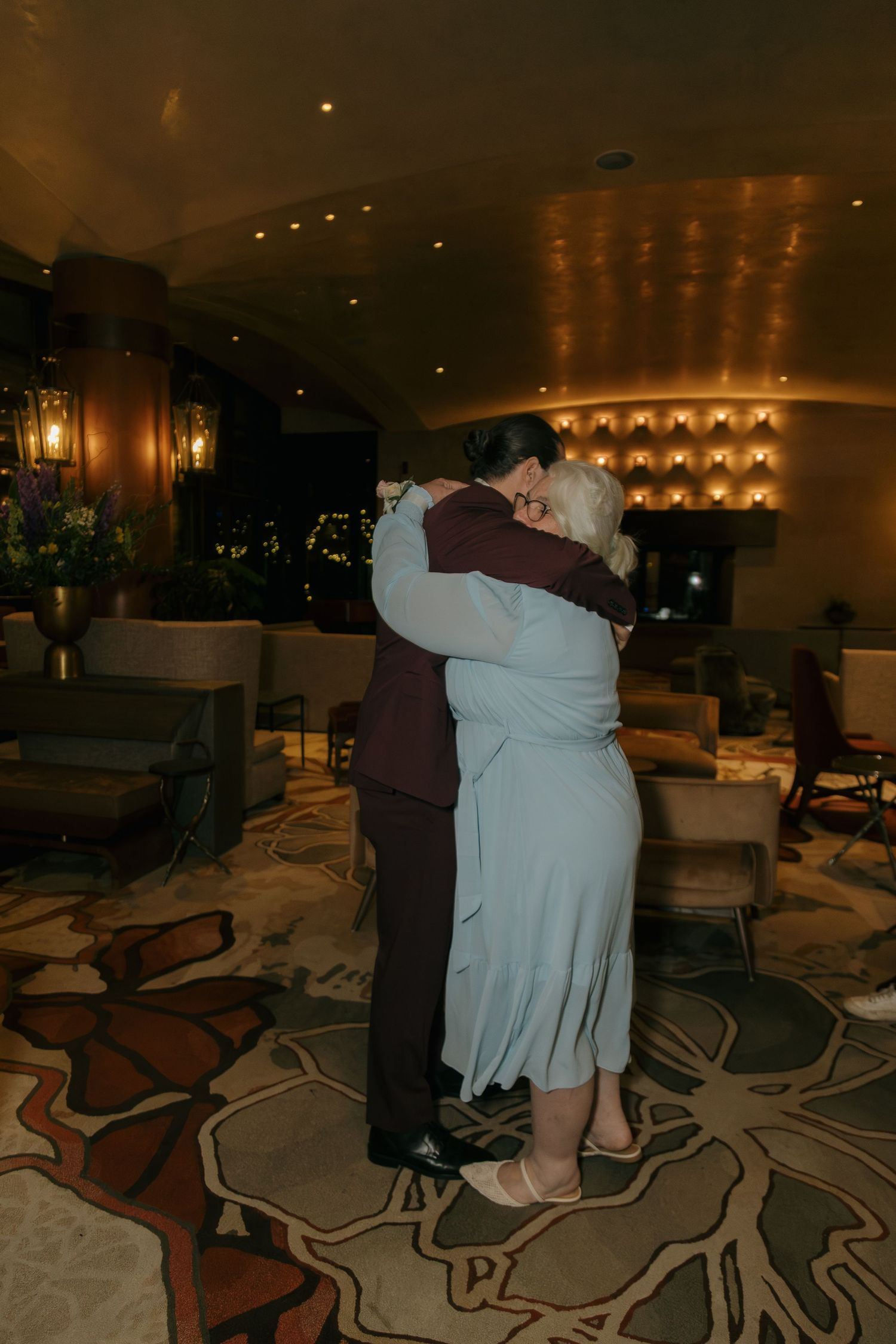 Two people share a warm embrace while dancing in an elegantly lit hotel lobby during a wedding reception.