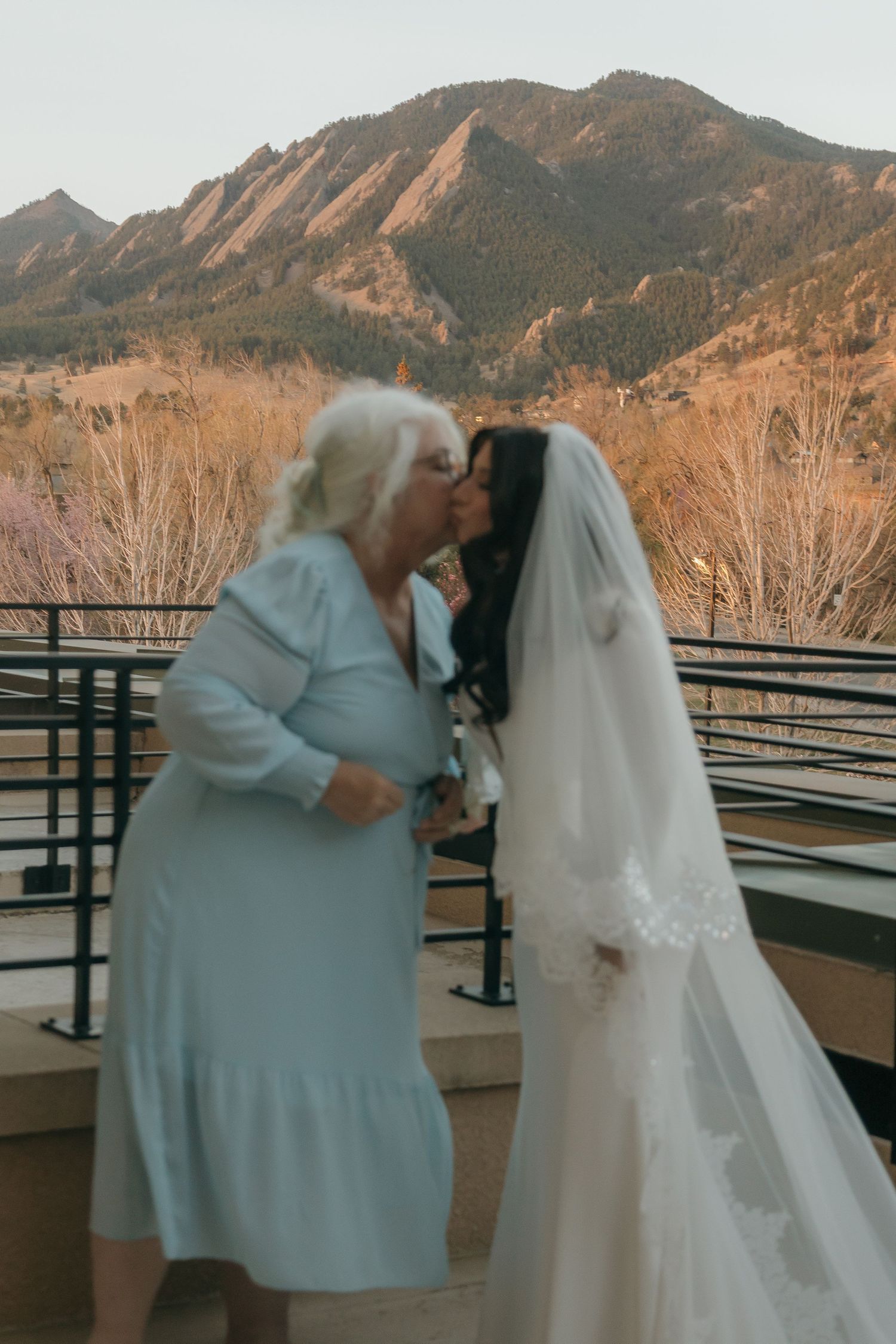 Bride in wedding gown kissing older woman with Flatirons mountain backdrop at sunset.