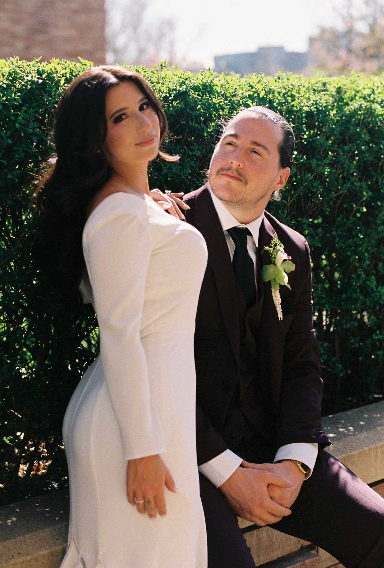 Bride in elegant white long-sleeve gown poses with groom in black suit beside lush green hedge.