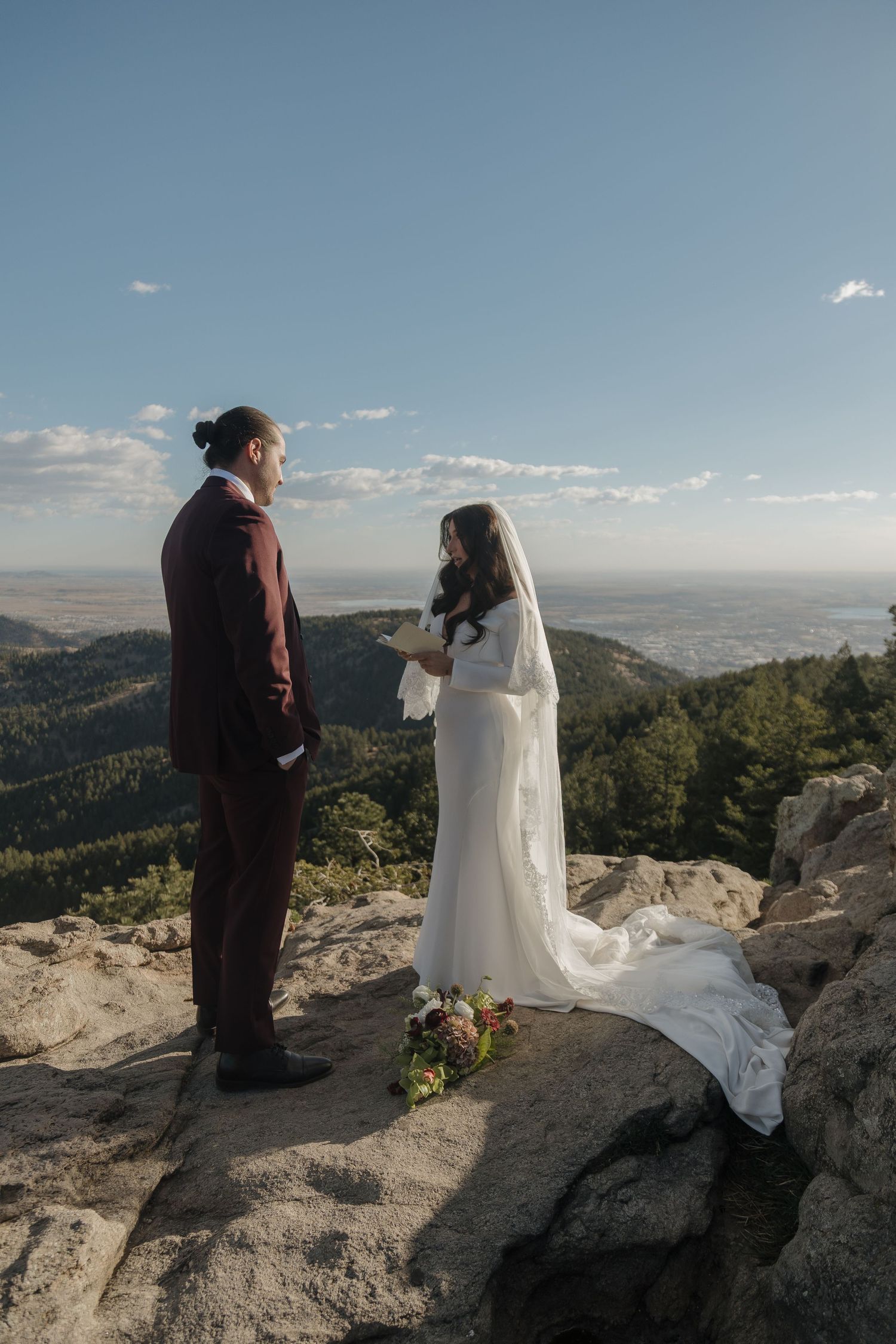 Bride and groom exchange vows on a rocky mountain summit with a sweeping valley view under a clear blue sky.