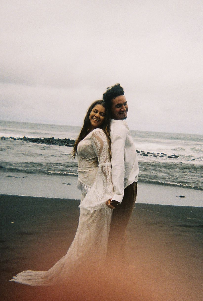 Joyful couple in wedding attire smile on a black sand beach with overcast ocean waves behind.