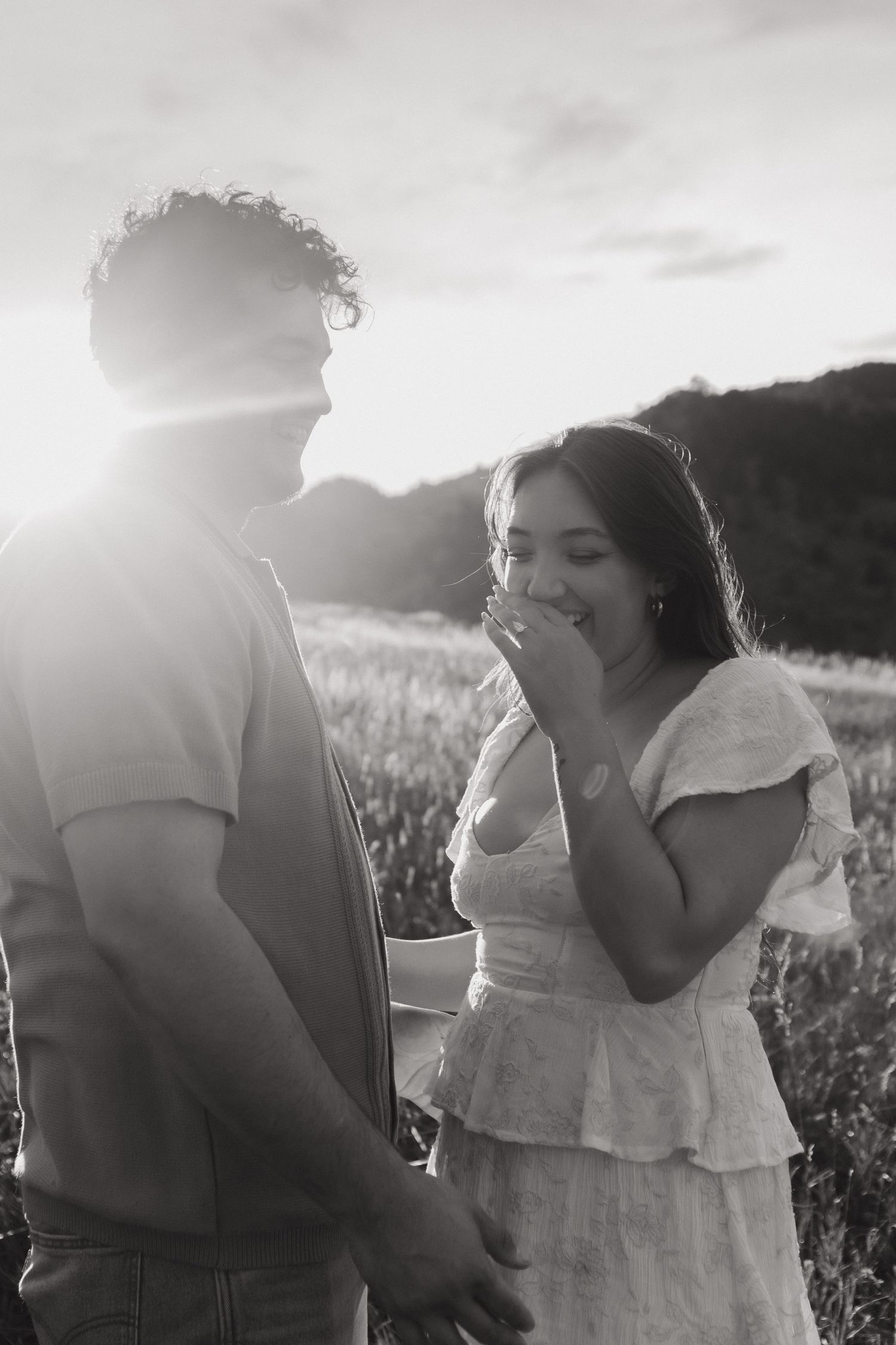 Black and white photo of couple laughing together in field during sunset engagement session.