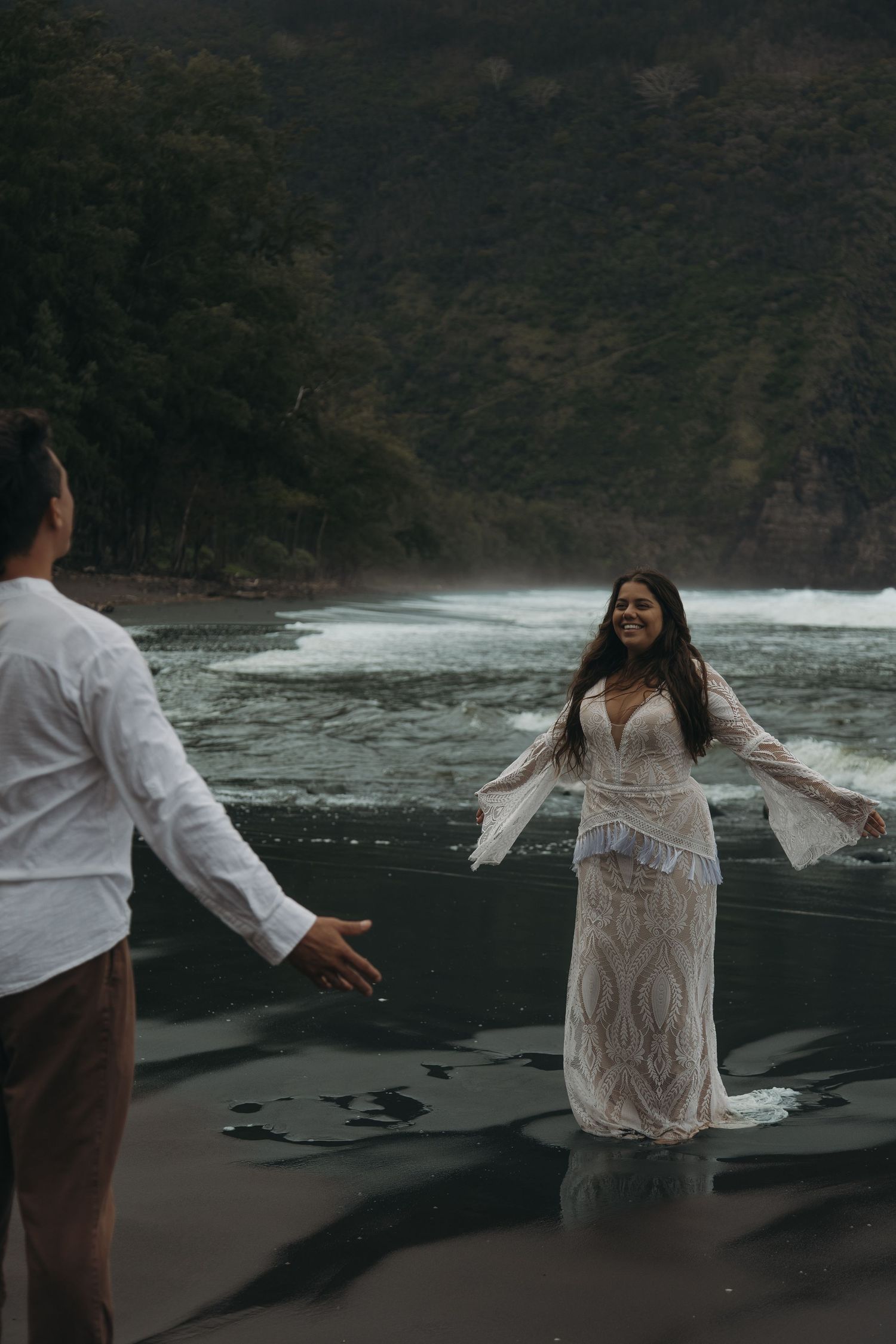 Smiling bride in white lace dress stands in ocean waves reaching toward groom on black sand beach.