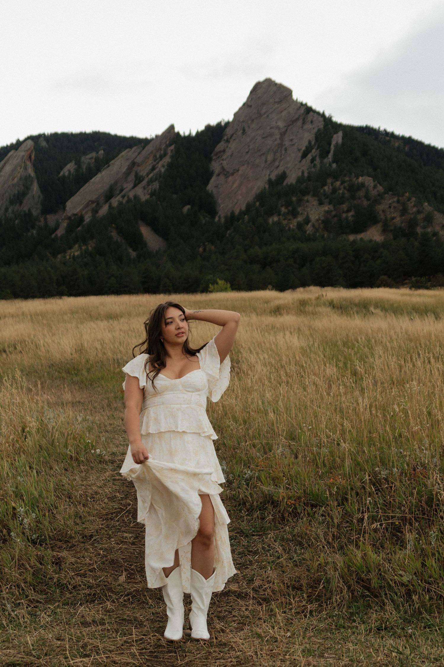 Woman in white tiered dress and cowboy boots poses in golden field with rocky Flatirons mountains backdrop.