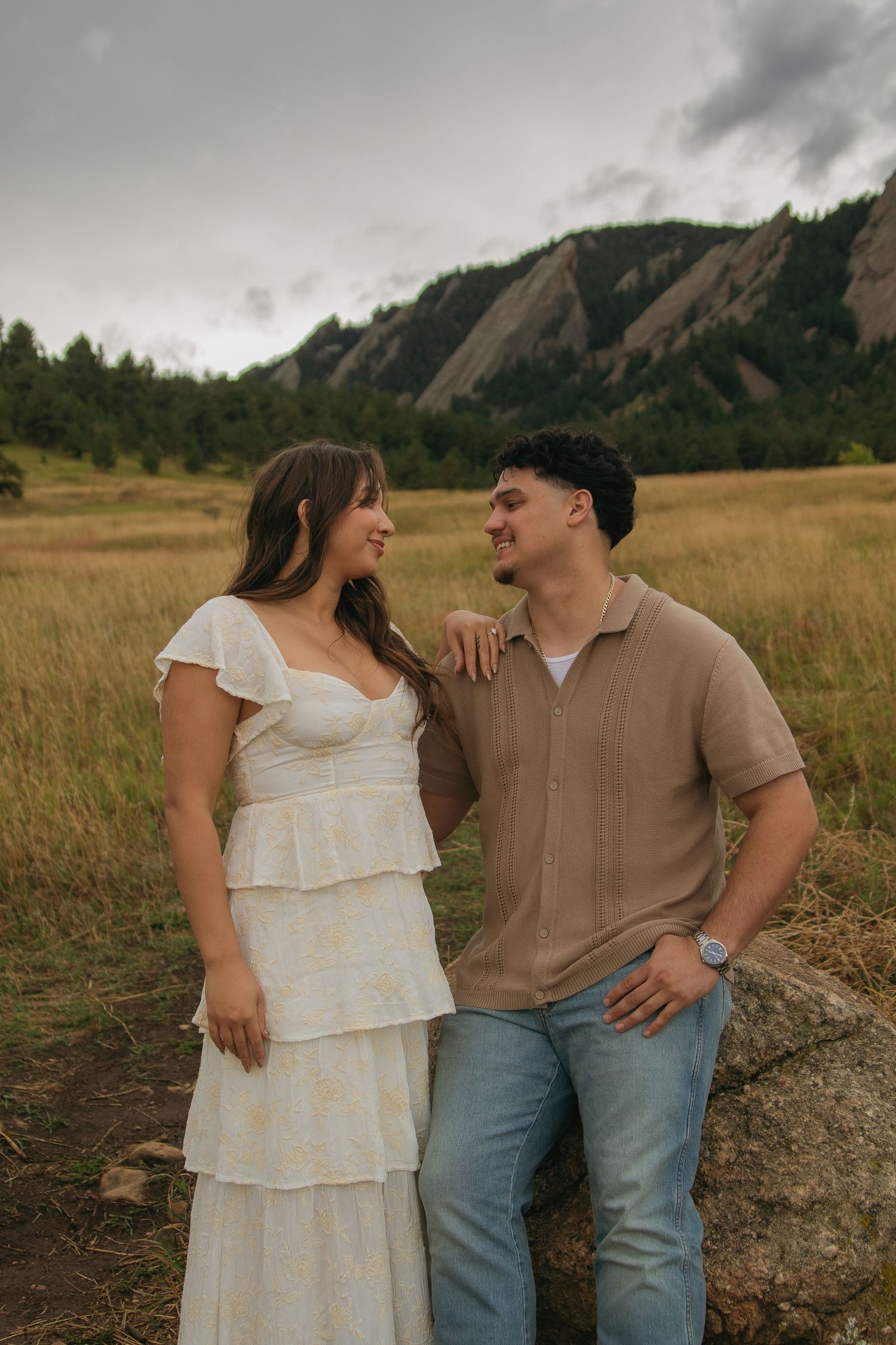 Smiling couple standing together in a field with the iconic Flatirons rock formations in background.