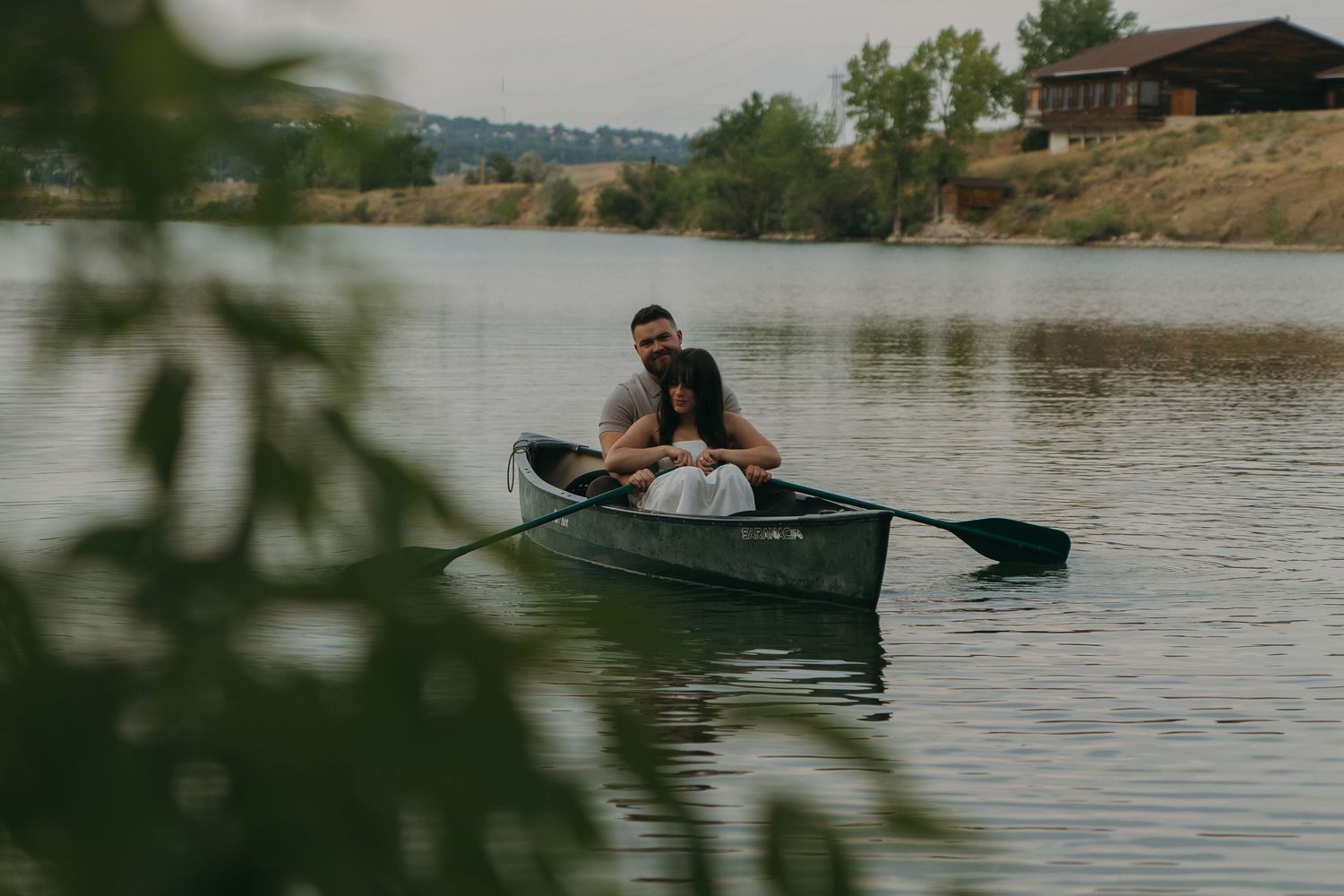 Couple sitting together in a canoe on a calm lake surrounded by lush green trees and hills.