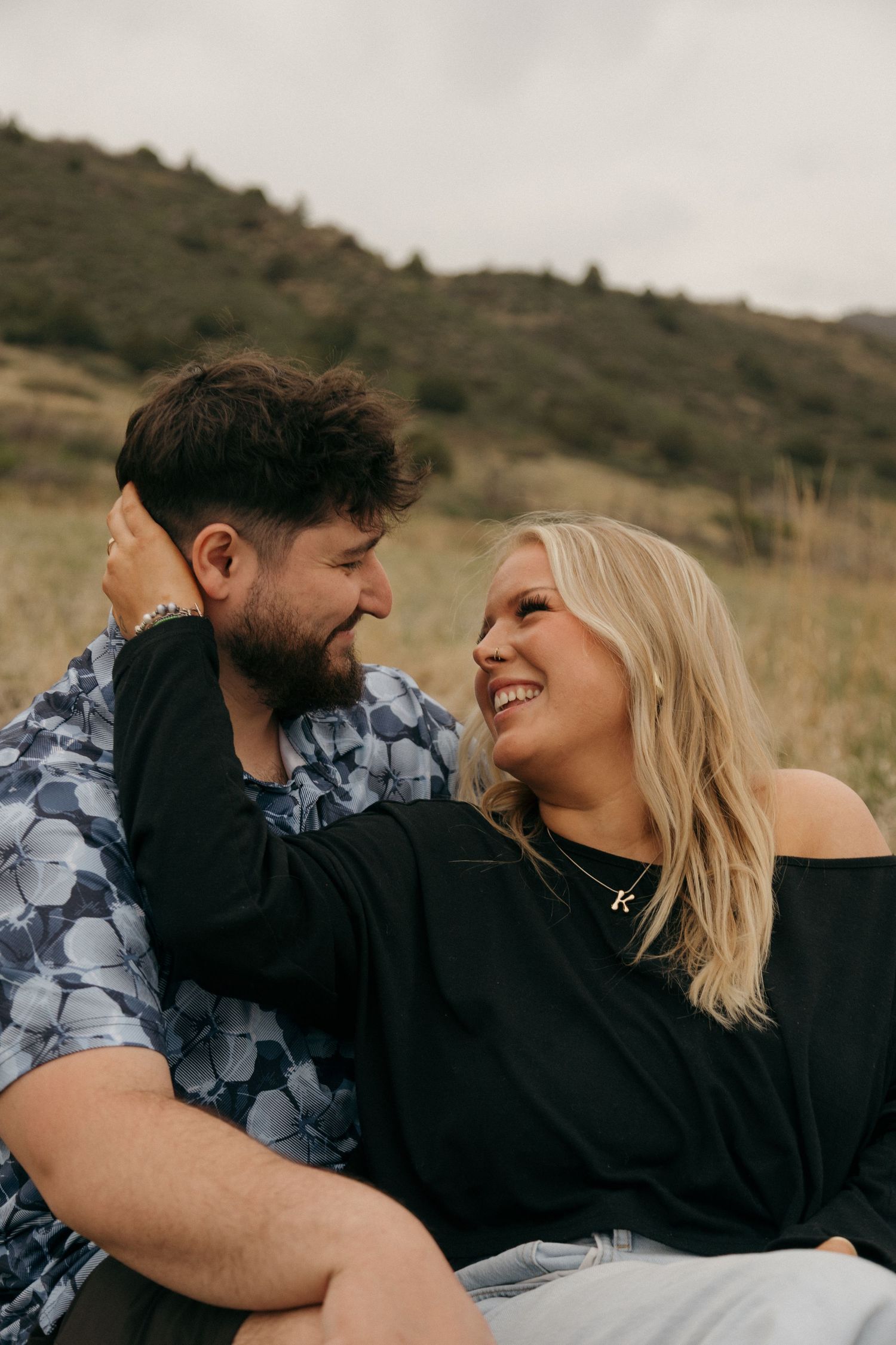 A smiling couple sits in a grassy field, the woman in black touching the bearded man's face as they gaze at each other.