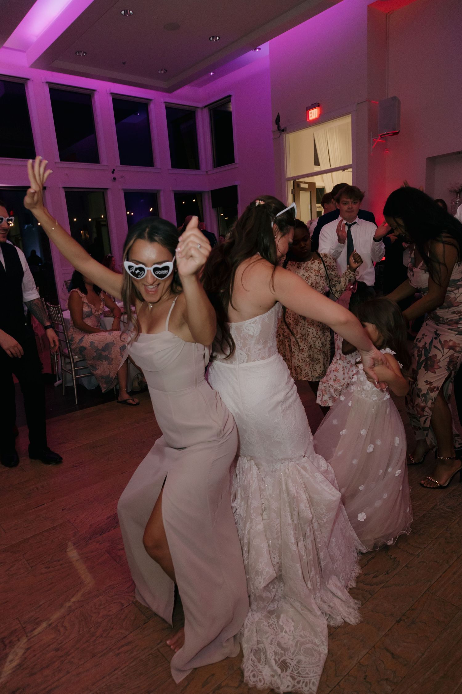 Two women in white dresses dancing energetically at a vibrant wedding reception with purple lighting.