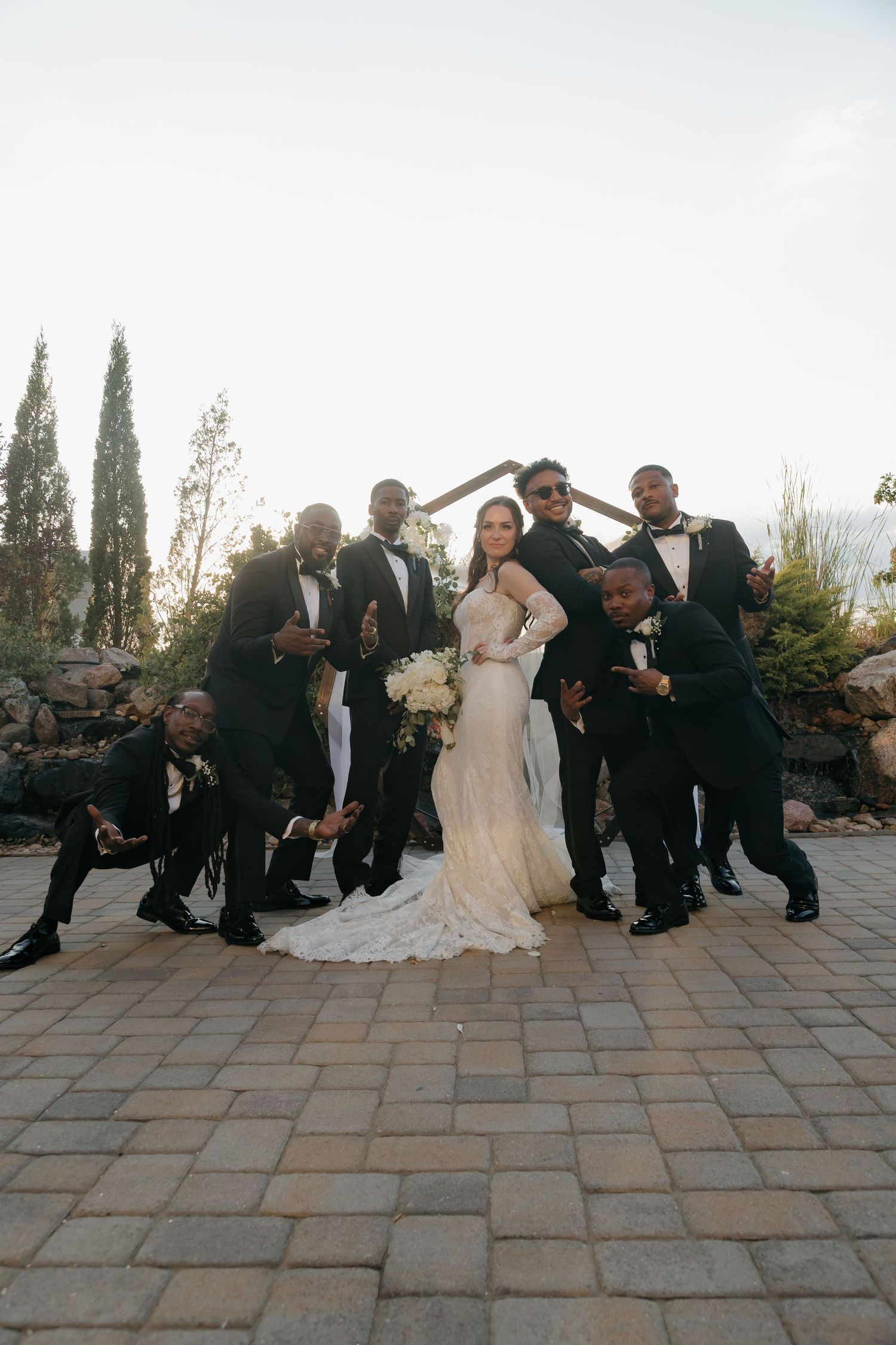 Bride in fitted lace gown poses with groomsmen in black tuxedos on stone patio at sunset wedding.